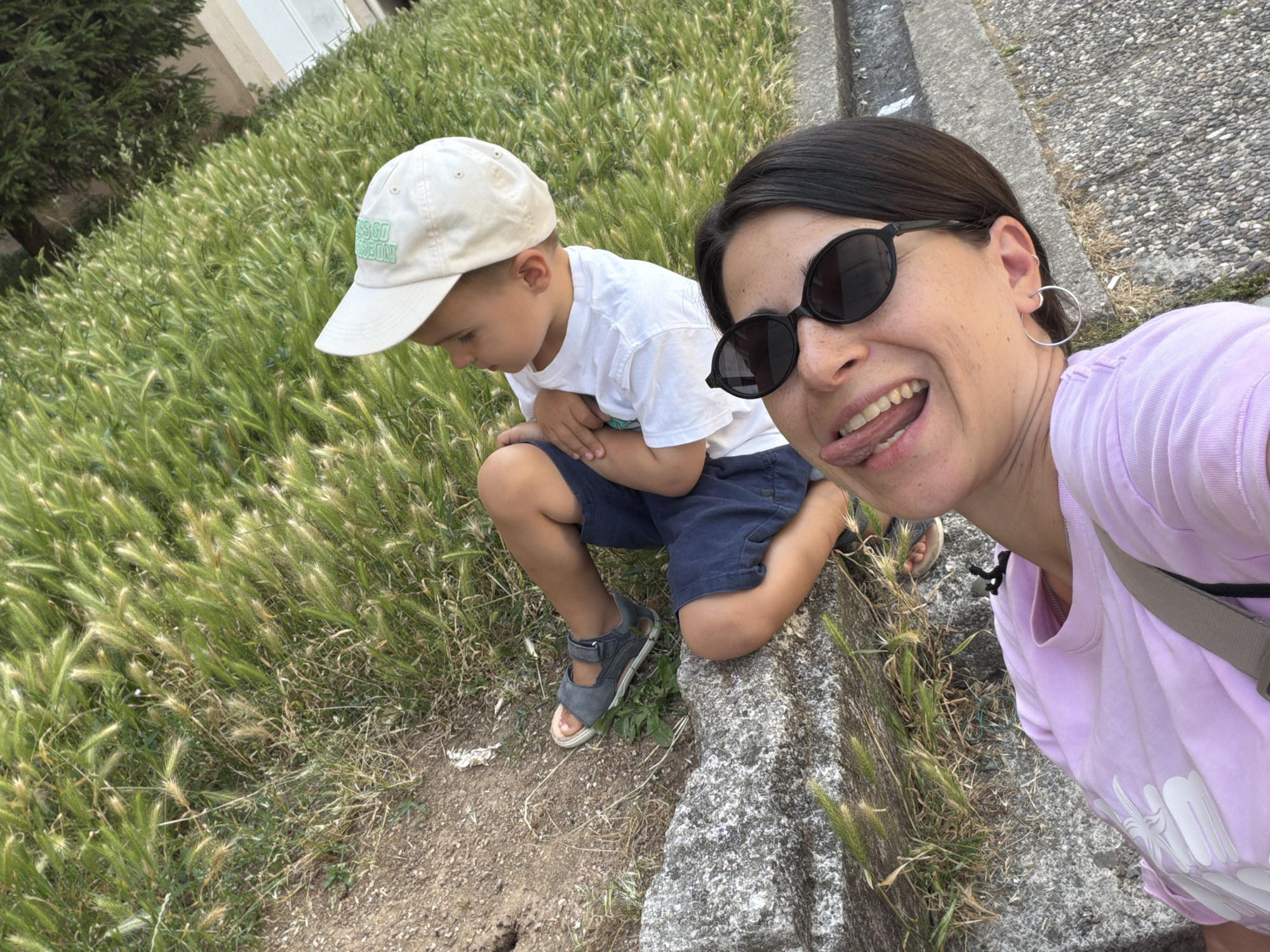 A woman takes a selfie with a child while sitting on a stone structure in Budva, Montenegro. The child, wearing a white t-shirt, blue shorts, and a white cap, is looking down. The woman, wearing sunglasses and a lavender t-shirt, has her tongue out in a playful manner. Both are seated near tall grass.
