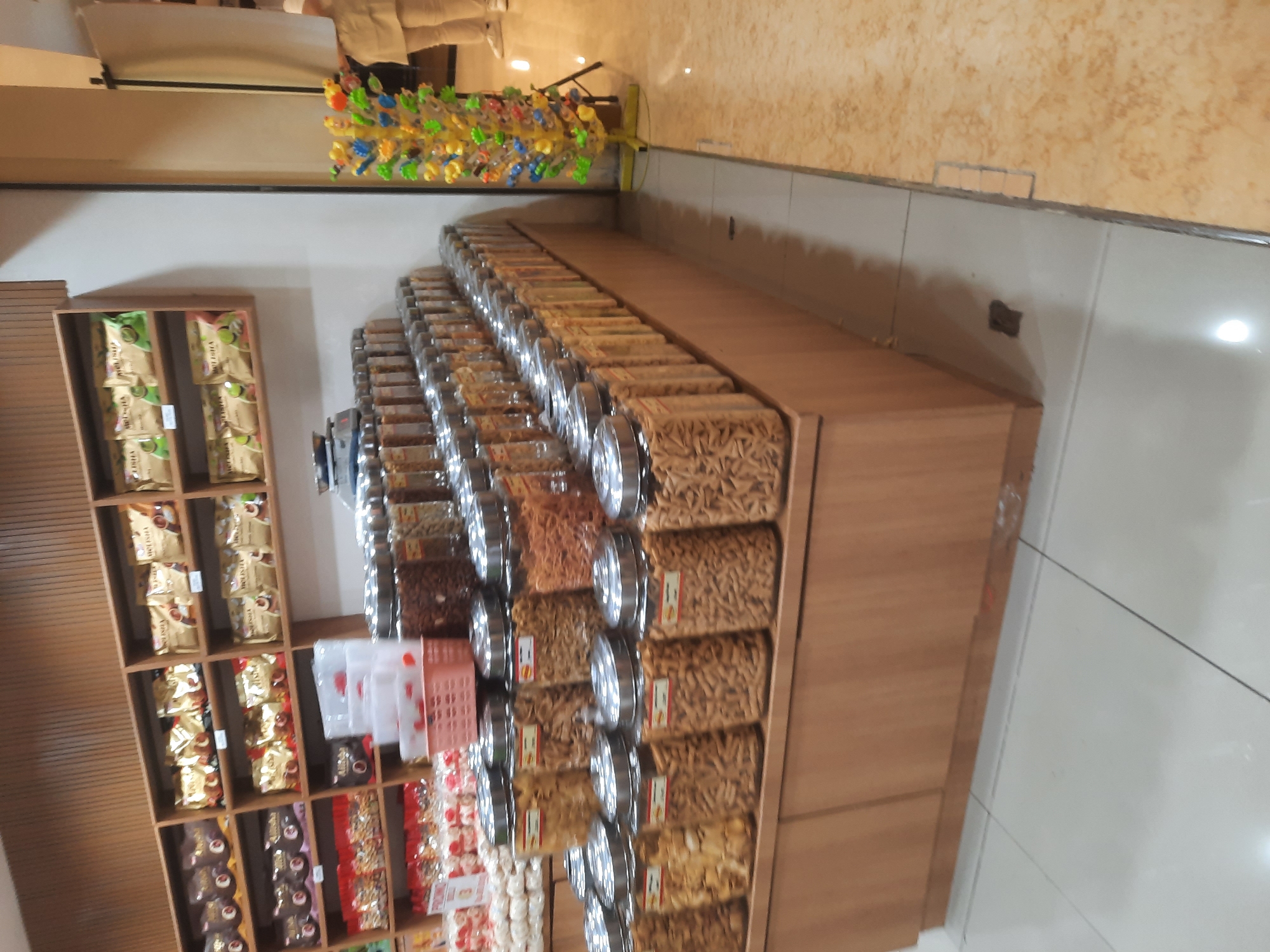 This is a medium-shot, eye-level photograph taken in a store in North Jakarta, Indonesia. The scene depicts a display of packaged snacks and bulk snacks in jars. The snacks are arranged on wooden shelves and a counter. The shelves in the background are filled with bags of snacks, many of which appear to be gold-colored with brown lettering. In the foreground, a tiered display of clear plastic jars filled with various types of snacks is prominent. These jars have silver lids. There is also a pink plastic basket filled with what appear to be small snack bags or portions. In the far right of the image, a person wearing a light-colored uniform and white shoes is partially visible, standing near a colorful display of small toys on a stand. The floor is tiled with a light gray and beige marble pattern. The overall environment suggests a retail setting, likely a snack or specialty food store.