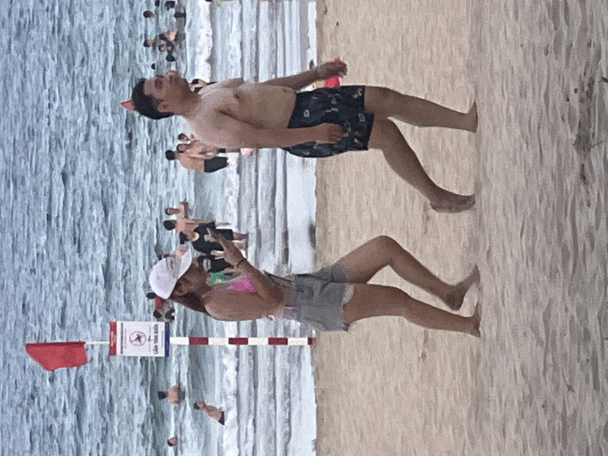 A man and a woman are walking on a sandy beach in Da Nang, Vietnam. The man, in the foreground, is shirtless and wearing dark patterned swim trunks. The woman, to his left and slightly behind him, is wearing a pink bikini top, grey denim shorts, and a white baseball cap. She is looking down at her phone. In the background, the ocean is visible with several people swimming. A red flag is flying on a pole near the water's edge, and a sign with a symbol of a person swimming in rough water is posted on the pole. The lighting suggests it is daytime.
