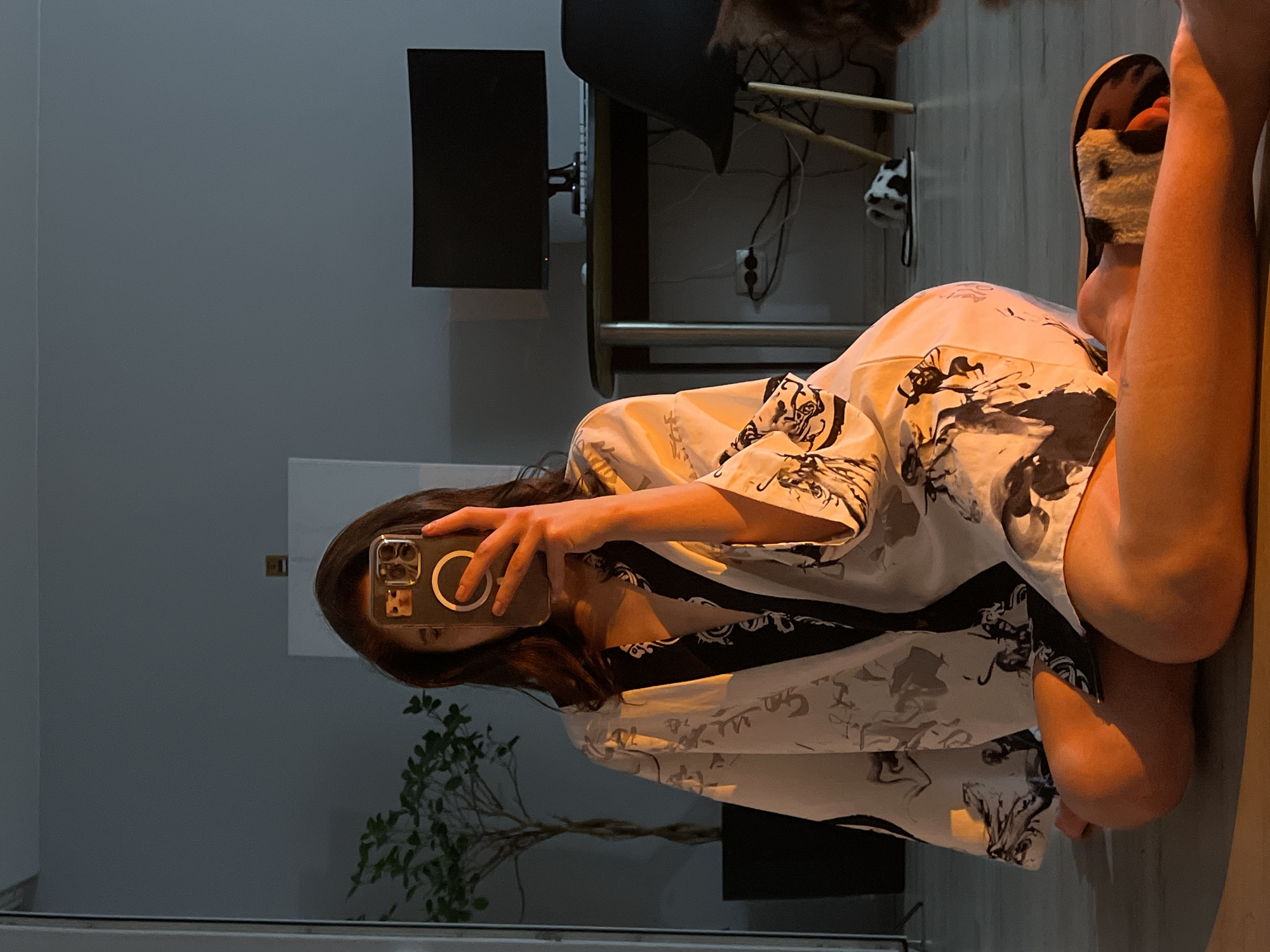 A woman is sitting on the floor in a dimly lit room, taking a selfie with her phone. She is wearing a light-colored robe with a black and white pattern. The room appears to be a home, with a desk, computer monitor, and chair in the background. A potted plant is visible to the left. The lighting suggests it might be evening, with a warm glow illuminating the woman. The location is not specified as Ternopil, Ukraine in the image.