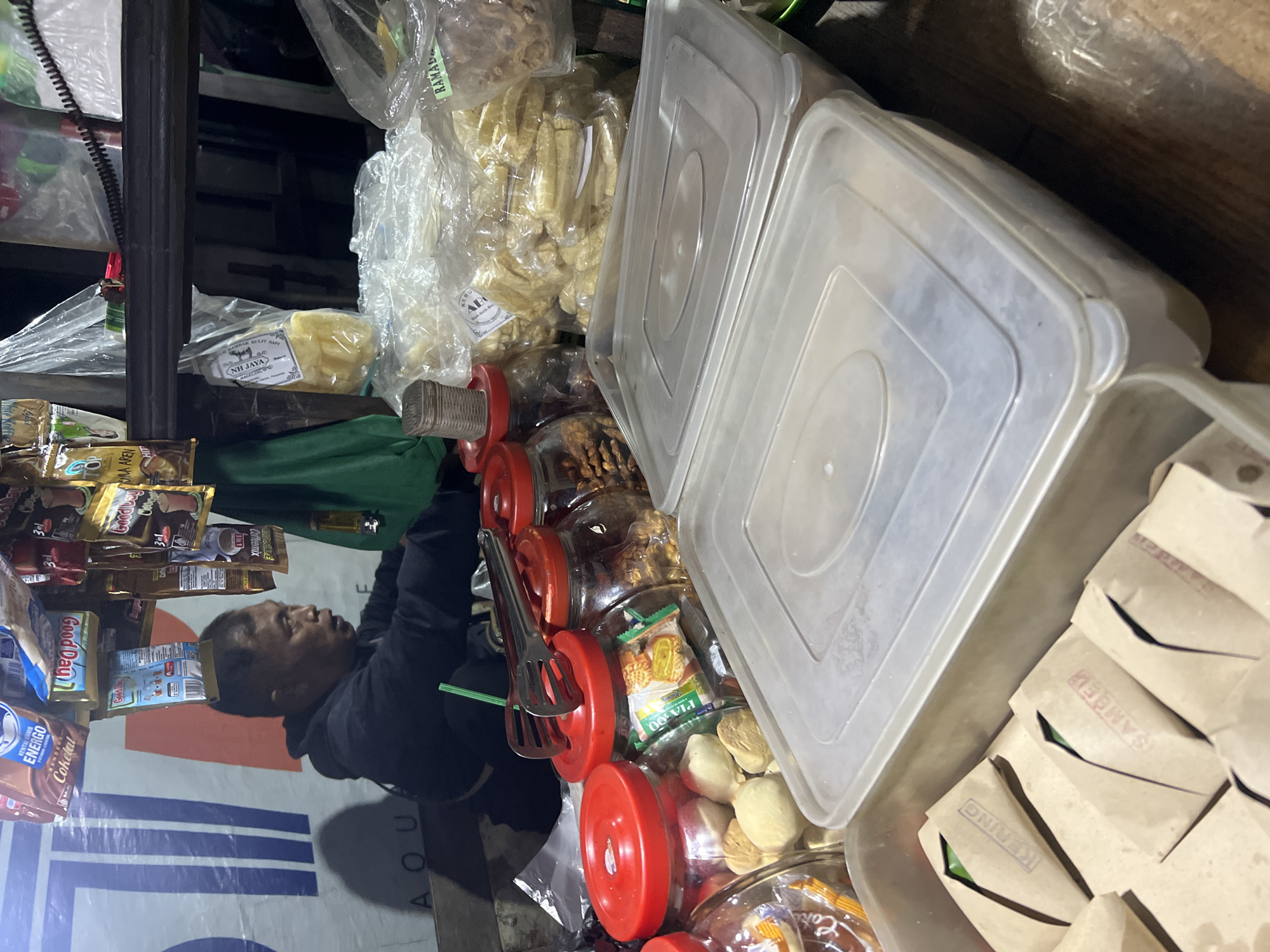 The image depicts a street vendor in Secang, Indonesia, operating a stall likely selling snacks and packaged goods. A middle-aged man, presumably the vendor, is visible from the chest up, looking to his right. He is wearing a dark hooded jacket. The stall is stocked with an assortment of items, including various packaged food products hanging from above, such as instant coffee and biscuits. In the foreground, there are clear plastic containers filled with what appear to be snacks like crackers and nuts. Below these, a long, shallow plastic container holds multiple small, individually wrapped packages made of brown paper, some bearing stamped labels. Behind the vendor, a green tarp is visible. The overall environment suggests an outdoor nighttime or dimly lit setting, typical of a street market.