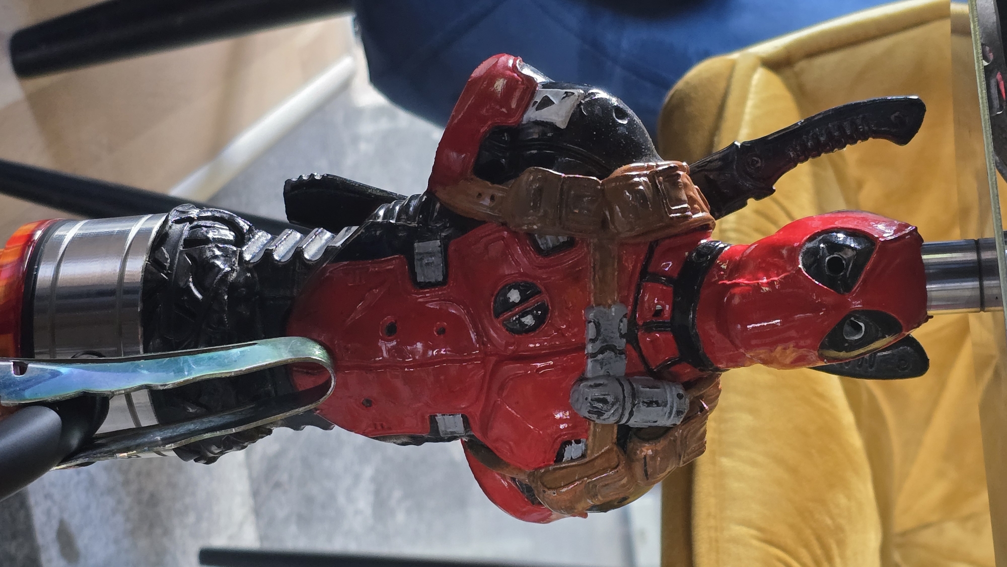 A close-up shot of a Deadpool figure on what appears to be a vaping device, laying on its side. The figure is predominantly red and black with brown straps and grey detailing. The vaping device components are chrome and black, with a section near the figure's feet appearing to be translucent and iridescent. The background features a blurred yellow cushion and a dark object above, suggesting an indoor setting. The image does not provide sufficient information to determine its location in Sofia, Bulgaria.