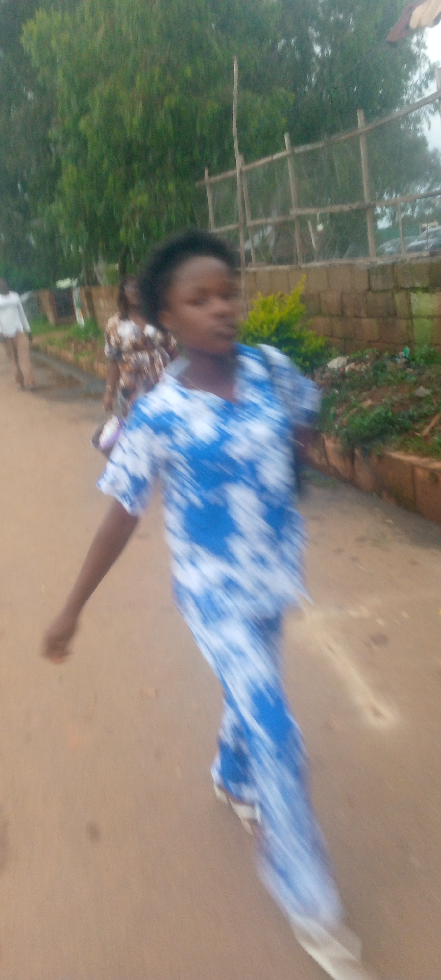 In an outdoor, urban environment, a young woman with dark hair is captured in motion, walking from left to right across a dirt or paved pathway. She is wearing a blue and white tie-dye pattern garment that appears to be a dress or a matching set of top and pants. Her right arm is extended out to her side. Behind her, another person is visible, also walking, wearing a patterned dress. Further in the background, a person in a light-colored shirt and pants is walking away from the viewer. The background includes lush green trees, a wooden fence structure, and a brick wall. The overall lighting suggests an overcast or late afternoon setting.