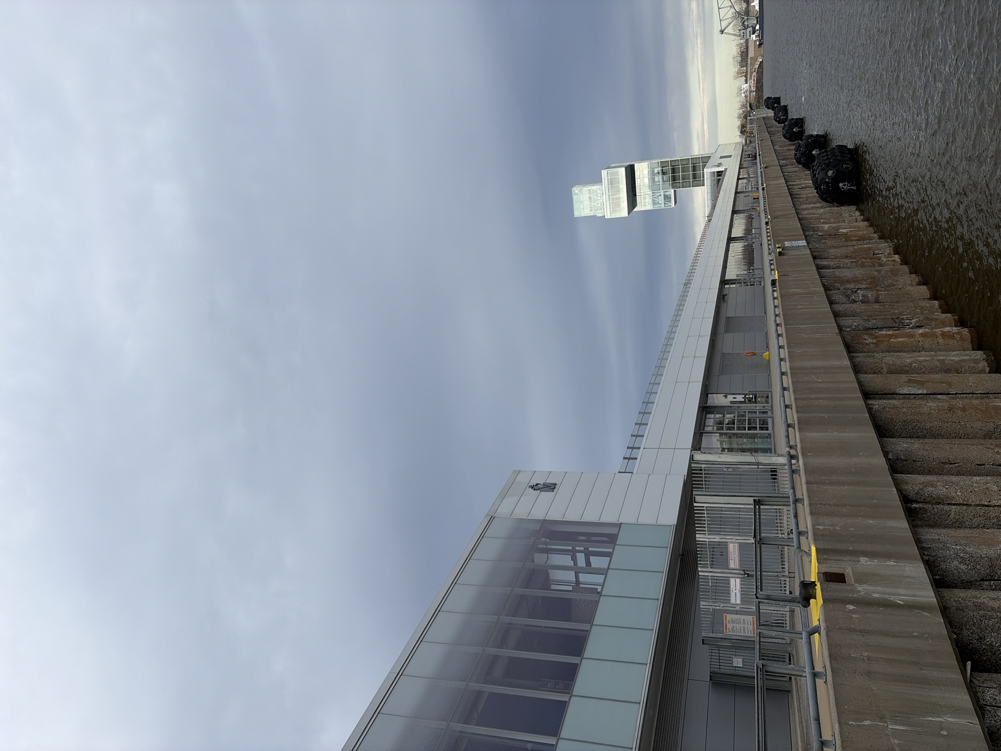 The image shows the exterior of a modern building in Montreal, Canada, located along the water's edge. The building features a combination of glass and white paneling, with a stacked cube design rising vertically. Next to the building is a concrete embankment. The water is visible to the right of the embankment, with a row of black buoys floating on its surface. The sky is overcast.