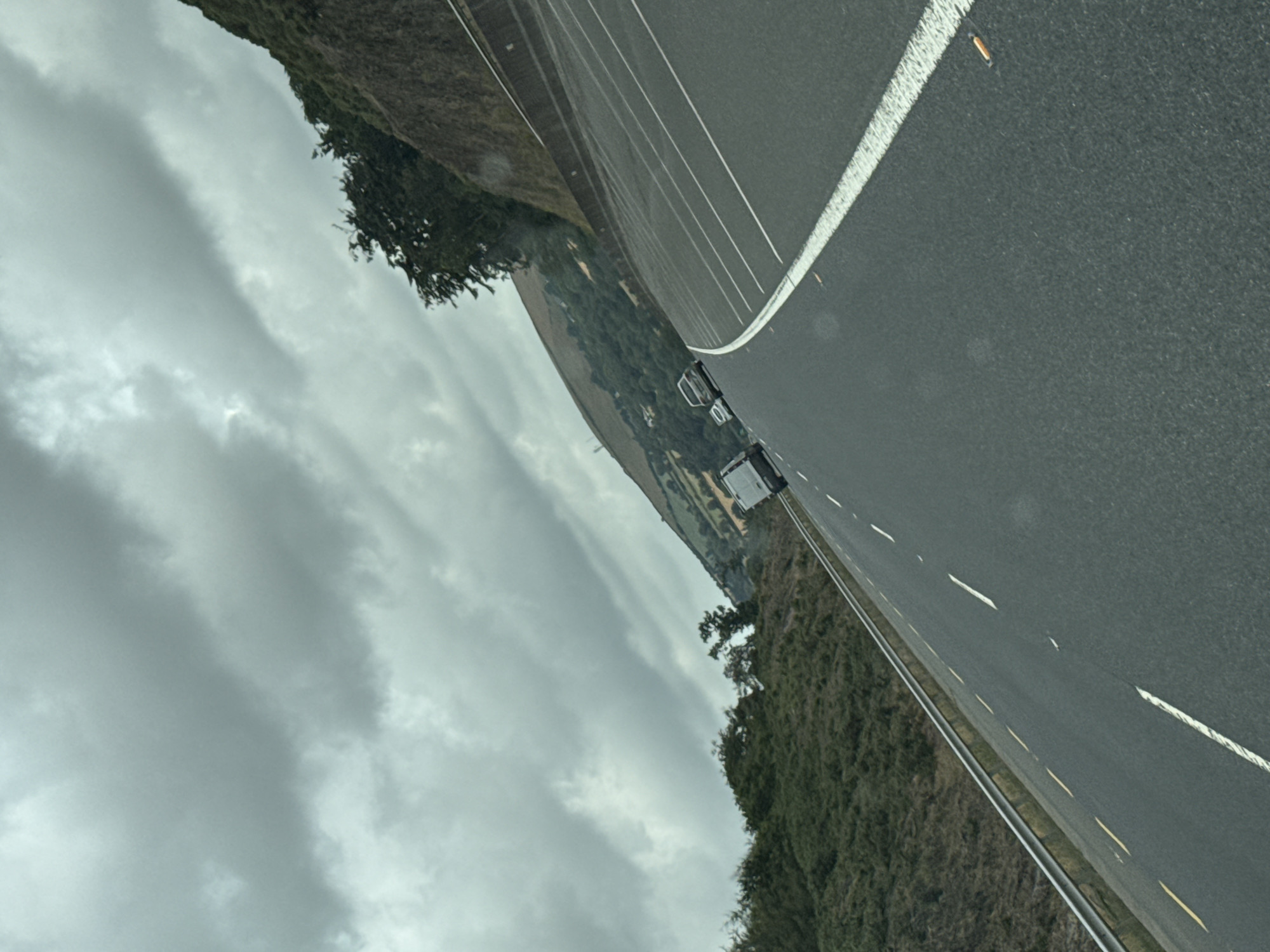A white van and two cars are driving on a wet asphalt road with white lane markings. The road is bordered by a metal guardrail on the left and a steep, vegetated embankment on the right. The sky is overcast with dark, heavy clouds. The scene appears to be a highway or major road in a rural or semi-rural area. The image is tilted, suggesting it was taken from a moving vehicle. The location is likely County Wexford, Ireland, possibly on the N25 road near New Ross, given the commonality of such landscapes in the region.