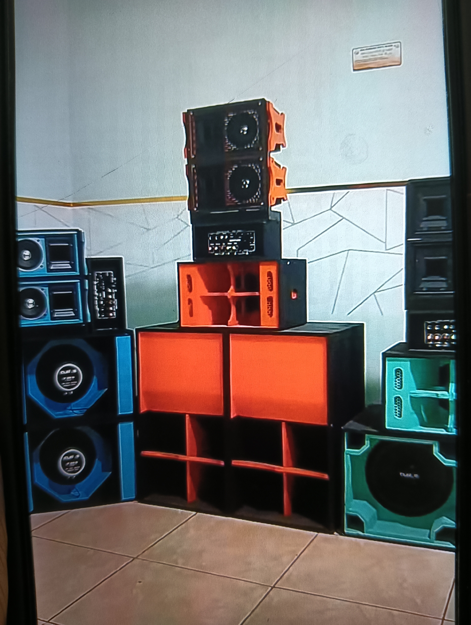 This is a photograph of a sound system setup in what appears to be an indoor space, possibly a room or a garage, with tiled flooring and a white wall featuring a geometric pattern. The sound system consists of multiple large speaker cabinets stacked in a complex arrangement.
On the left side, there are two blue speaker cabinets stacked vertically. The top cabinet has two smaller circular speakers and two rectangular components. The bottom blue cabinet is larger and has a prominent circular speaker with a logo or text in the center. To the right of the blue speakers, there is a tall, narrow black speaker cabinet with multiple small circular elements on its face, also stacked on top of another black cabinet.
In the center of the image, there are several orange speaker cabinets. The top orange cabinet is a dual-speaker unit with two circular speakers. Below this are two orange cabinets side-by-side, each appearing to have internal shelving or dividers.
On the right side of the image, there are black speaker cabinets stacked vertically. The topmost black cabinet has visible components resembling speakers or other audio equipment. Below this are two turquoise-colored speaker cabinets stacked. The bottom turquoise cabinet is a large enclosure with a prominent circular speaker.
A small, rectangular orange-and-white sign is mounted on the upper right portion of the wall. The overall impression is that of a powerful audio setup. There are no people visible in the image. The location in the city of Semen, Indonesia, is not visually apparent from the provided image.