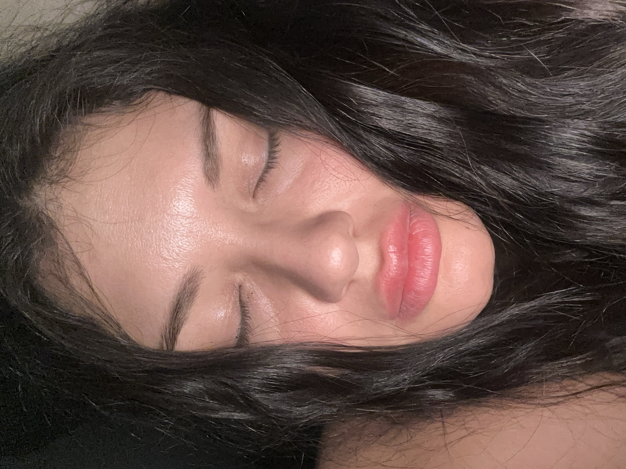 A close-up image captures the face of a woman with dark, wavy hair surrounding her face. Her eyes are closed, and she appears to be sleeping. Her skin has a natural, slightly glossy sheen. Her lips are pink and full. The lighting is subdued, creating a soft effect. There is no indication that the image was taken in Khimki, Russia.