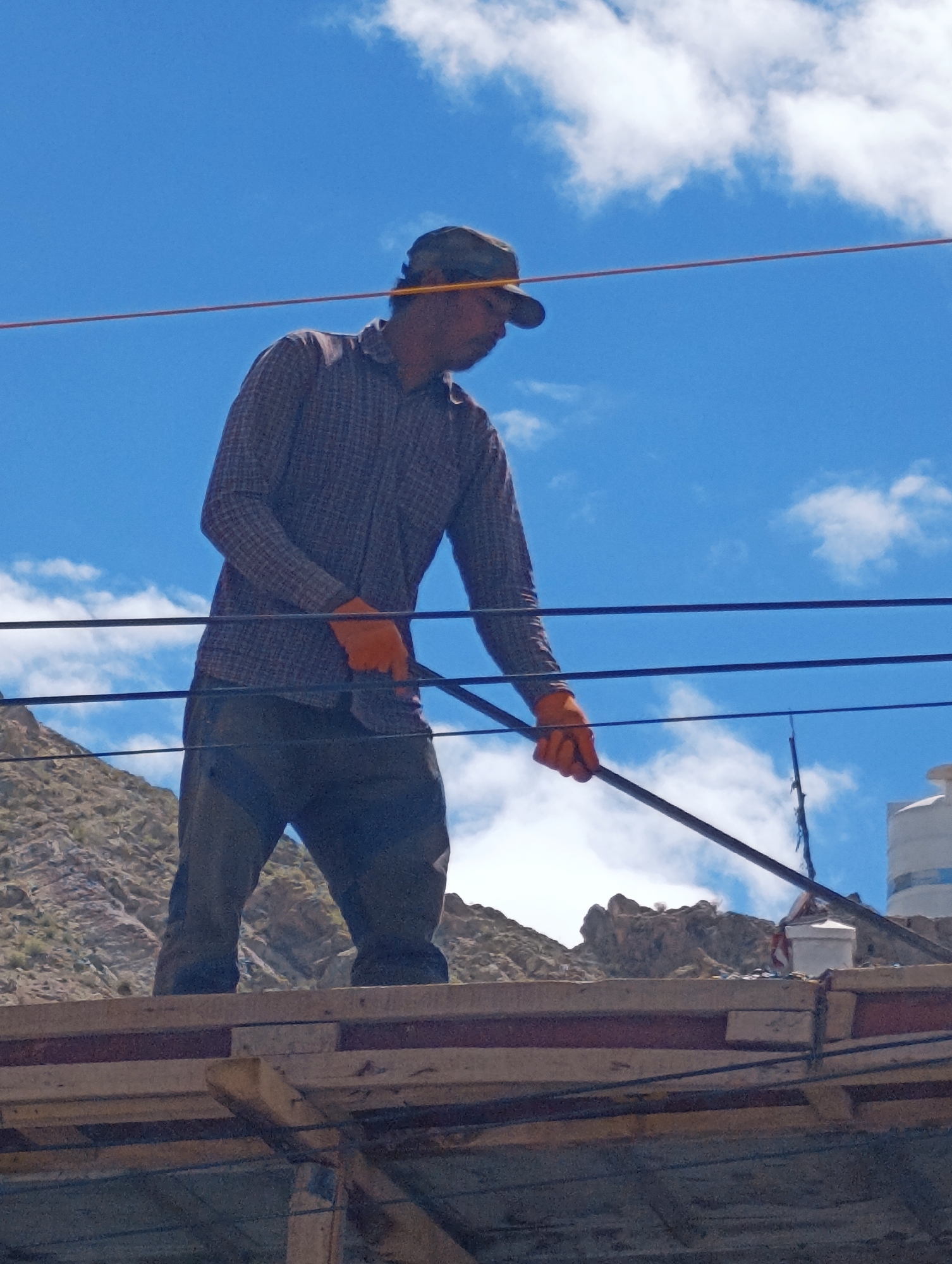 A man wearing a checkered shirt, dark pants, a cap, and orange gloves stands on a wooden structure under a bright blue sky with scattered white clouds. He is holding a long pole horizontally with both hands, angled downwards and to the right. In the background, a rocky, barren mountain slope is visible, with some structures, including a white water tank, on a rooftop. The scene appears to be outdoors, possibly in a construction or work area. The location is identified as the city of Leh, India.