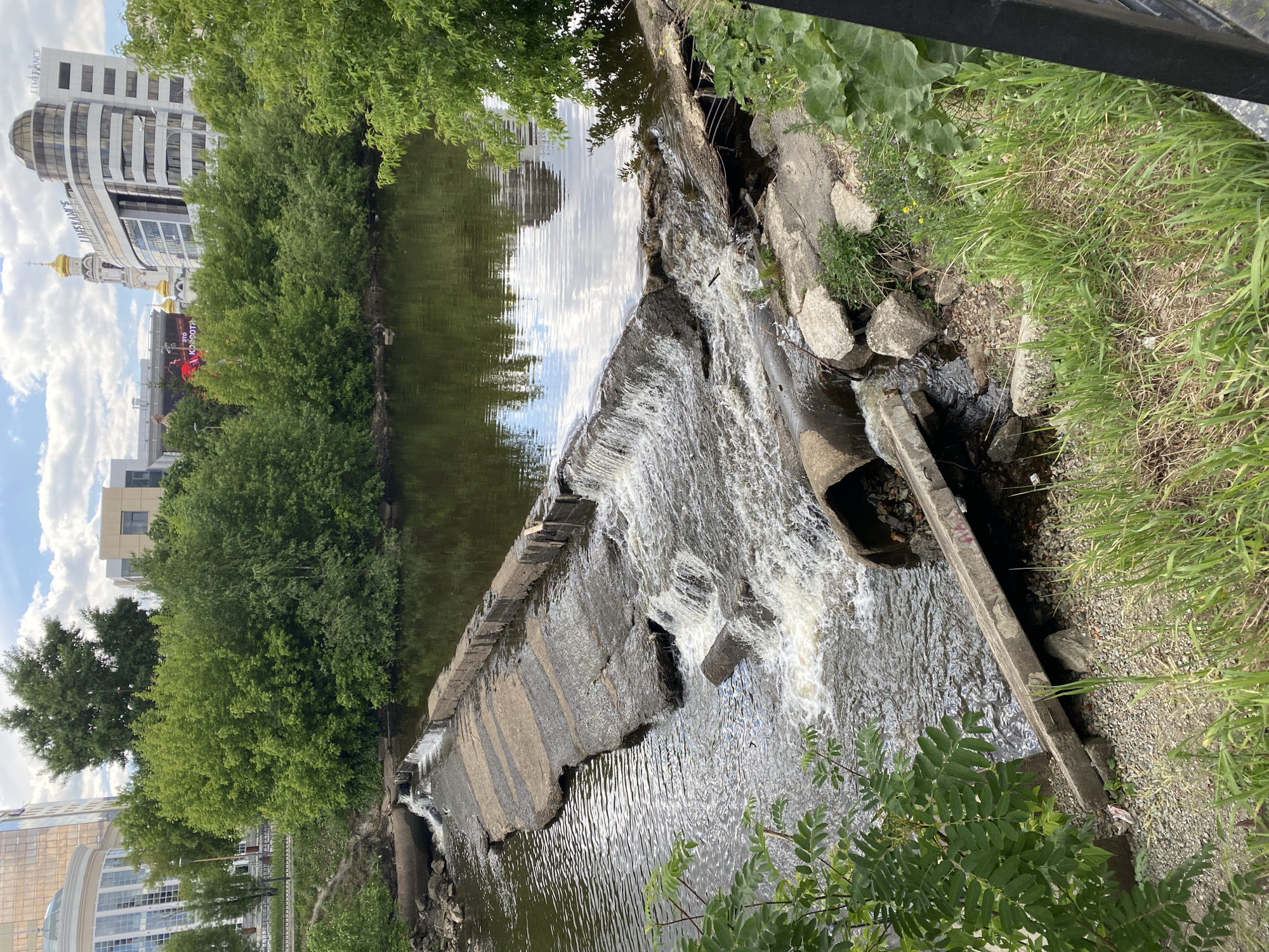 This image depicts a man-made waterfall in the city of Yekaterinburg, Russia. The waterfall is created by a series of concrete structures that span a river or canal. The water cascades over these structures, creating rapids and turbulence. Lush green vegetation lines the banks of the waterway, and buildings are visible in the background, indicating an urban setting. The scene is captured from an elevated perspective, providing a clear view of the waterfall and its surroundings.