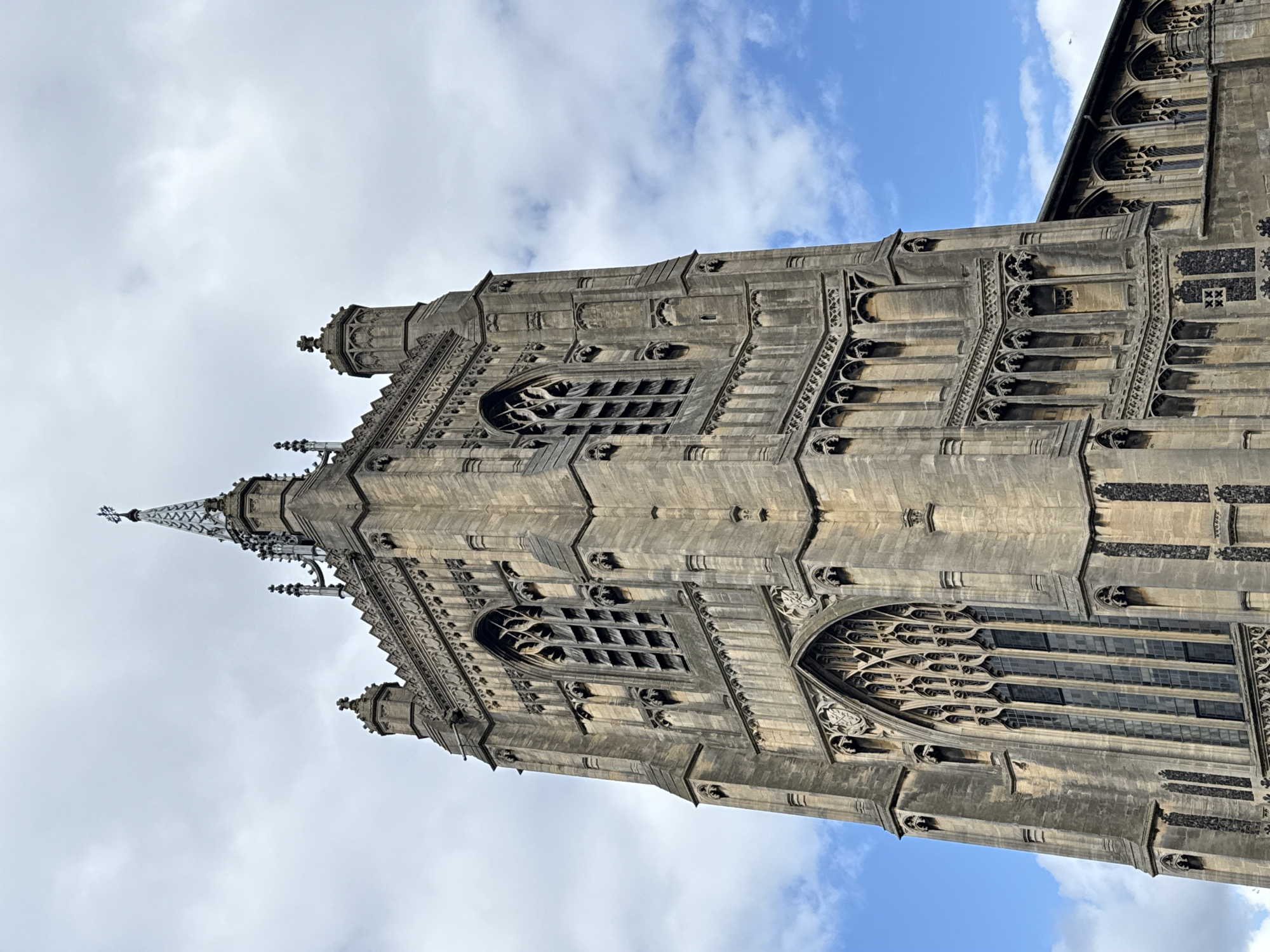 The image shows an upward view of the Perpendicular Gothic tower of Norwich Cathedral in Norwich, United Kingdom. The tower is made of stone and has several tiers of windows and decorative elements. The top of the tower is adorned with a spire. The sky is partly cloudy with patches of blue.