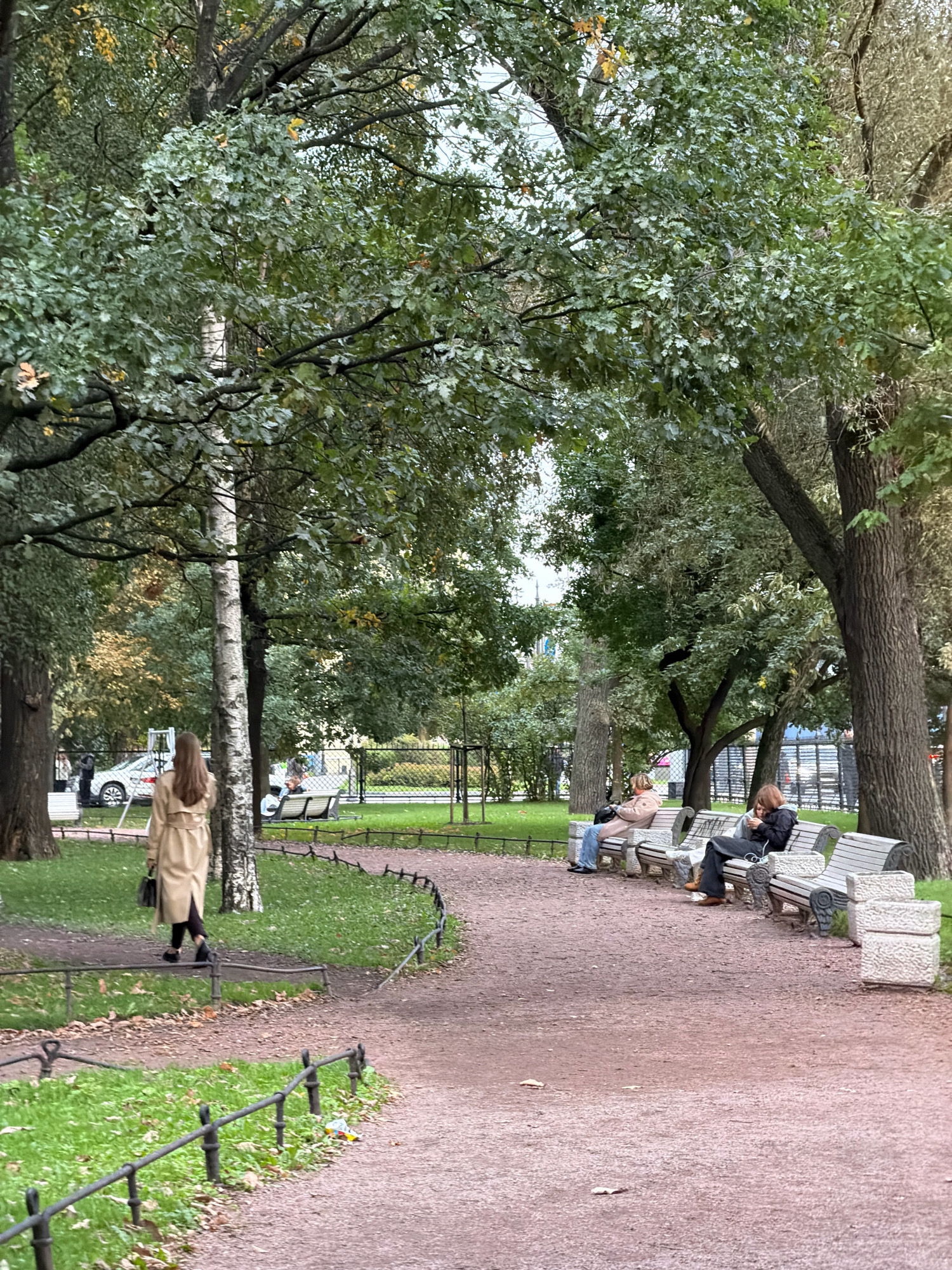 The image depicts a park scene in Saint Petersburg, Russia, likely during autumn due to the foliage colors. A woman in a beige trench coat walks away from the camera on a gravel path lined with a low, black metal fence. In the background, several people are seated on park benches. One person on a bench on the left wears blue jeans and a light-colored jacket. To the right, another person in a dark jacket is looking at their phone. The park features mature trees with dense green and some yellowing leaves, casting shadows on the path and grass. A white car is visible parked in the distance. The overall atmosphere is calm and leisurely.