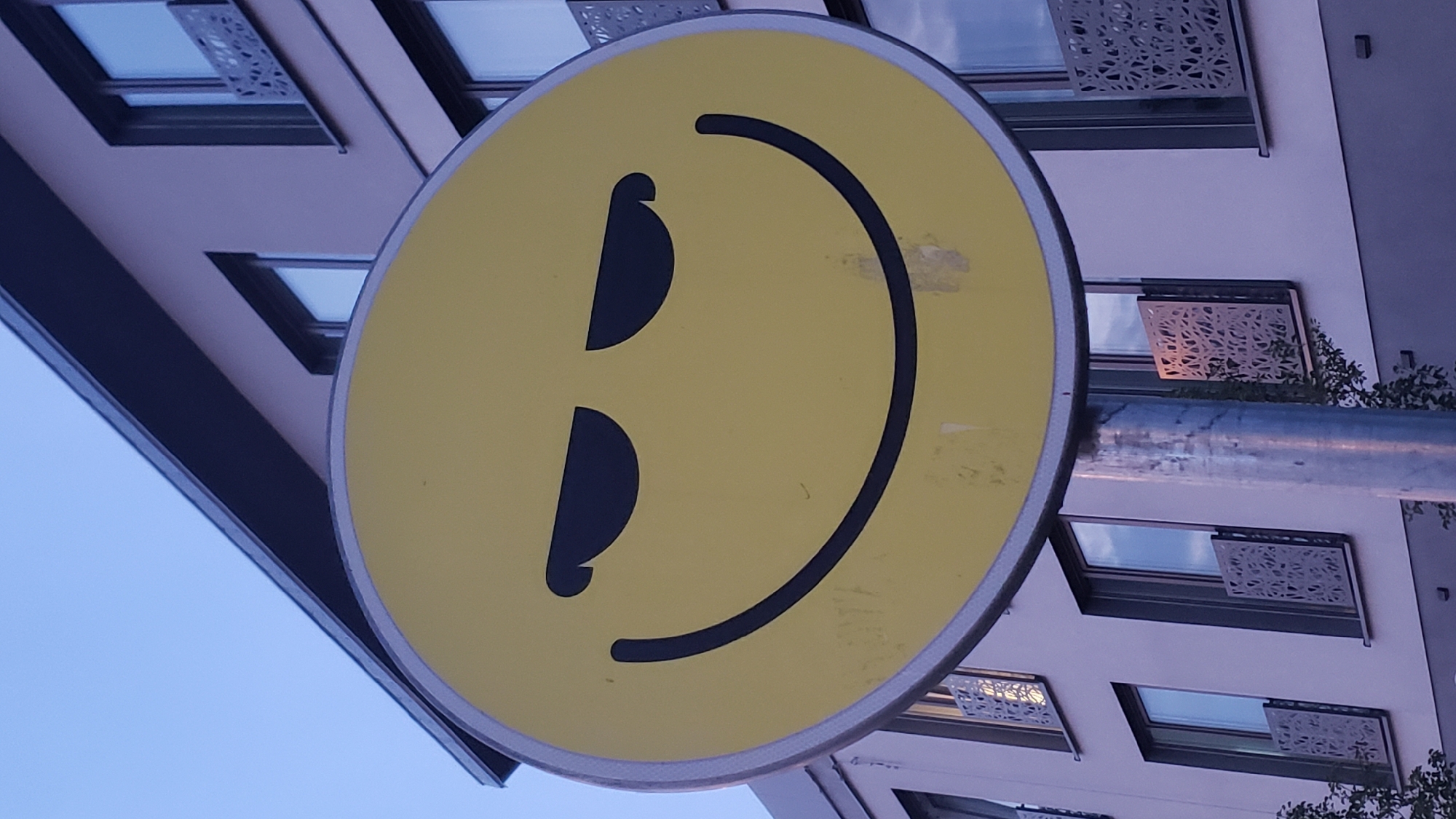 A yellow circular sign with a black smiley face is mounted on a metal pole in front of a modern apartment building. The smiley face has narrow, upward-slanted eyes, giving it a sly or mischievous expression. The building behind the sign is light-colored with multiple windows, some featuring decorative metal railings. The sky above is a pale blue. The scene appears to be an outdoor urban environment. The location of the image is the city of Prešov, Slovakia.