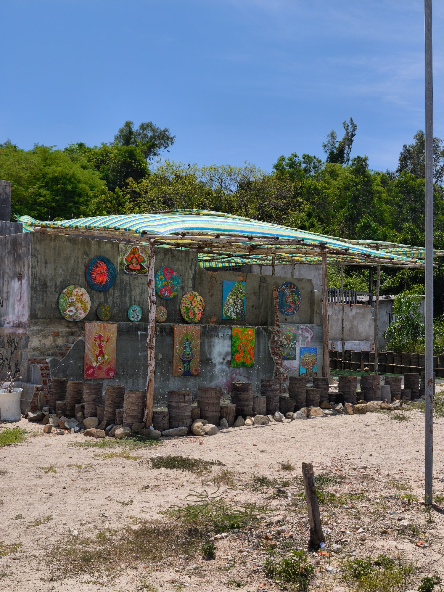 This is a wide shot of an outdoor art display against a backdrop of trees and a bright blue sky. The art is displayed on and around a rough, unfinished concrete structure covered by a striped green and yellow awning. The structure appears to be a lean-to or open-air shelter.
The concrete wall behind the display is adorned with numerous circular and rectangular paintings, depicting various colorful, often abstract or nature-inspired designs. Some of the circular paintings feature floral motifs and intricate patterns. Below the paintings, a row of roughly cut, cylindrical wooden posts, possibly tree trunks, are stacked and arranged. Rocks and broken bricks are scattered at the base of these posts.
To the left of the structure, a white pot with a small plant sits near the corner of the concrete wall. The ground in front of the display is sandy and somewhat uneven, with patches of dry grass and scattered debris. A tall metal pole stands to the right of the scene, extending from the foreground to the top of the frame. The overall impression is a rustic, open-air art exhibition, possibly in a coastal or rural setting near Vĩnh Lương, Vietnam, given the vegetation and the style of the display.