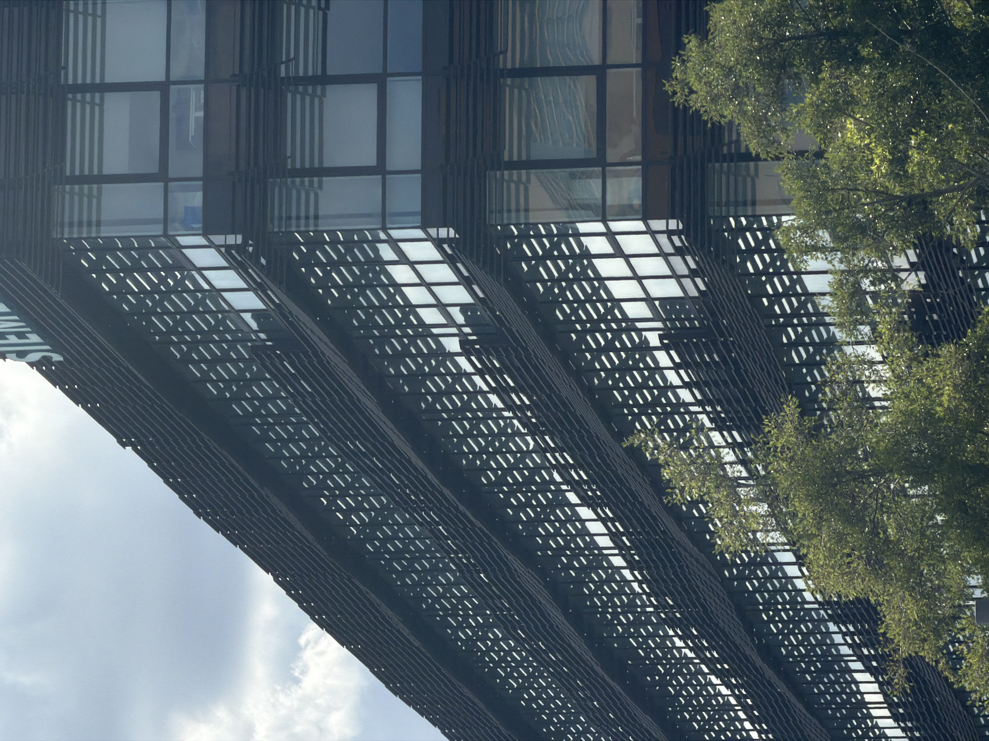 This image depicts a modern building with a distinctive facade, featuring a repeating pattern of horizontal louvers. The building's structure is characterized by dark metal framing and large glass windows, reflecting the sky. To the right of the building, lush green foliage from a tree cascades down, partially obscuring the architectural details. The sky is bright and partly cloudy, suggesting it is daytime with fair weather. The composition is angled, creating a sense of looking up at the building from below and to the side. The repeating patterns of the facade and the interaction with the natural element of the tree create a visually striking contrast between man-made design and nature. The scene appears to be an urban or suburban environment, possibly a business district or a modern residential complex. No people or text are visible in the image. The overall impression is one of contemporary architecture integrated with greenery.