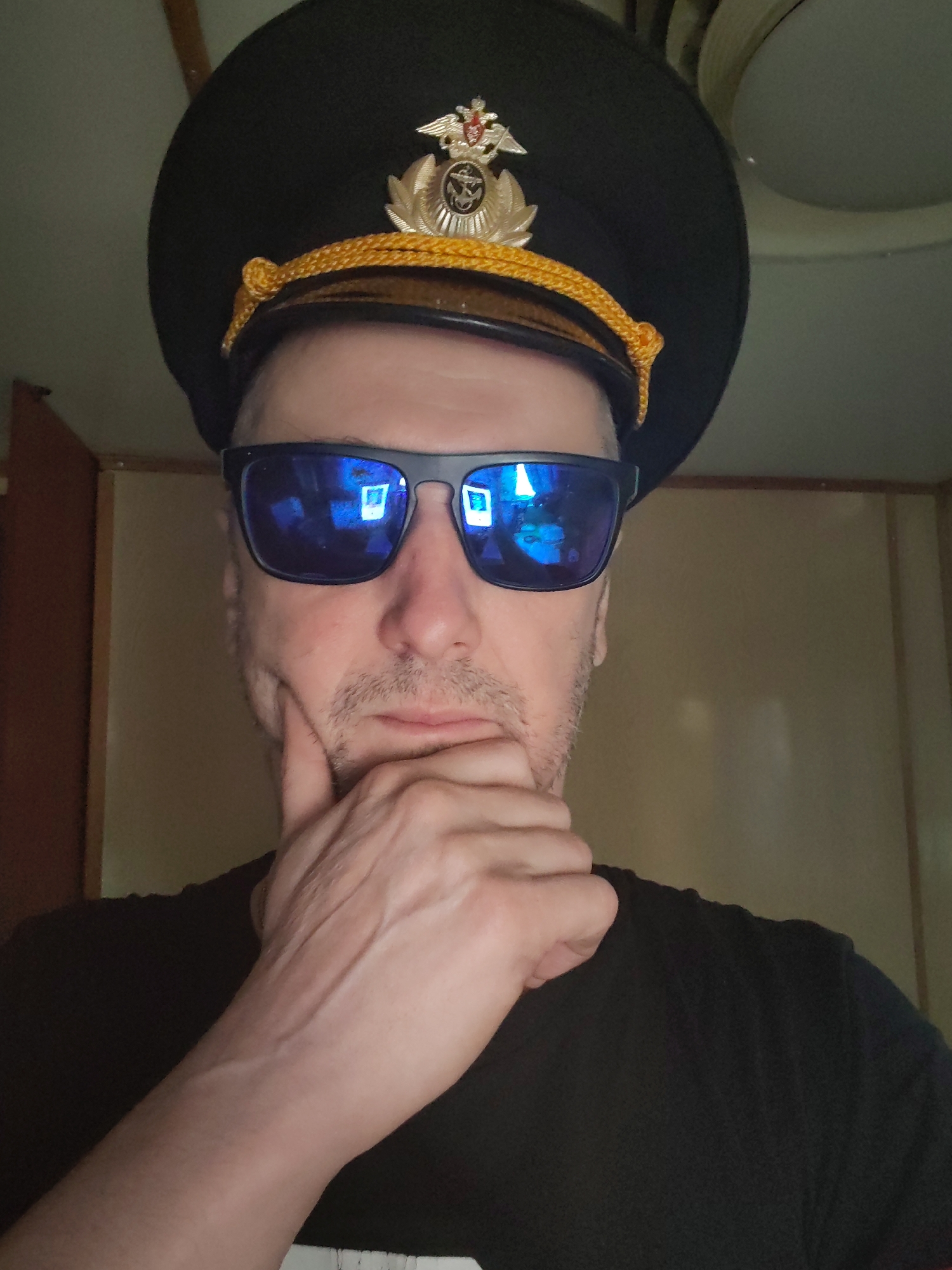 A close-up, front-facing portrait depicts a man indoors, likely in Shcheleiki, Russia. He is wearing a dark, peaked cap featuring a gold braided cord and a prominent golden insignia on the front, which appears to be a Russian Federation emblem with a double-headed eagle and an anchor symbol, suggesting a naval or military connection. He also sports dark, rectangular sunglasses with reflective blue lenses, obscuring his eyes and showing reflections of square light sources. Stubble is visible on his lower face. The man wears a plain black t-shirt, and his right hand is raised to his chin, with fingers resting beneath it in a thoughtful or contemplative pose. The background reveals a light-colored ceiling fixture in the upper right and simple wall paneling on the left and right sides. The lighting suggests an indoor environment.