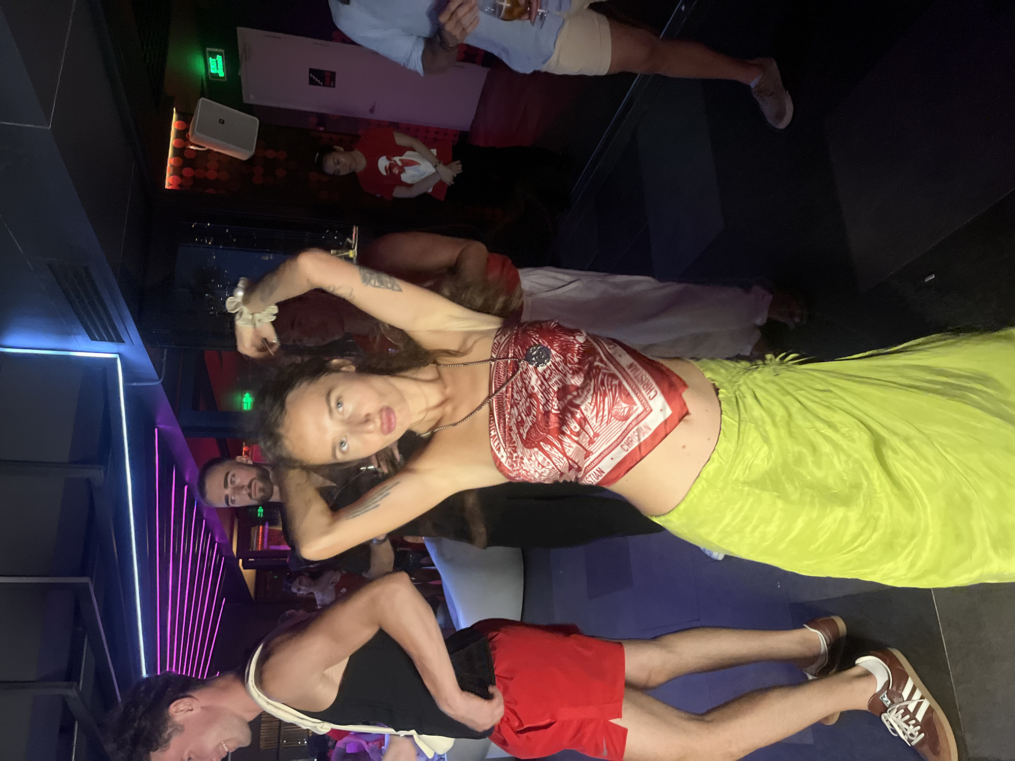 The image depicts a group of people at what appears to be a nightclub or entertainment venue. A woman in the foreground is the primary subject, wearing a red and white patterned scarf as a top and a bright green skirt. She is posing with her arms raised and her hands behind her head, looking directly at the camera with her tongue slightly out.
To her left is a man wearing a black tank top and red shorts, with a white strap over his shoulder. He is looking to his left and smiling. Behind him, another man with a beard is visible. Further in the background, a woman in a red t-shirt is standing, and on the far right, a man in a light blue shirt and beige shorts is holding a drink.
The environment is dimly lit, with neon purple lighting accents in the ceiling. There are speakers mounted on the wall and an "EXIT" sign illuminated in green. The floor is dark, with some reflective surfaces and what appear to be dark mats.
While the image is set in a nightclub, there is no specific visual information within the image to indicate that the location is in Đà Nẵng, Vietnam. The scene is typical of an indoor nightlife setting.