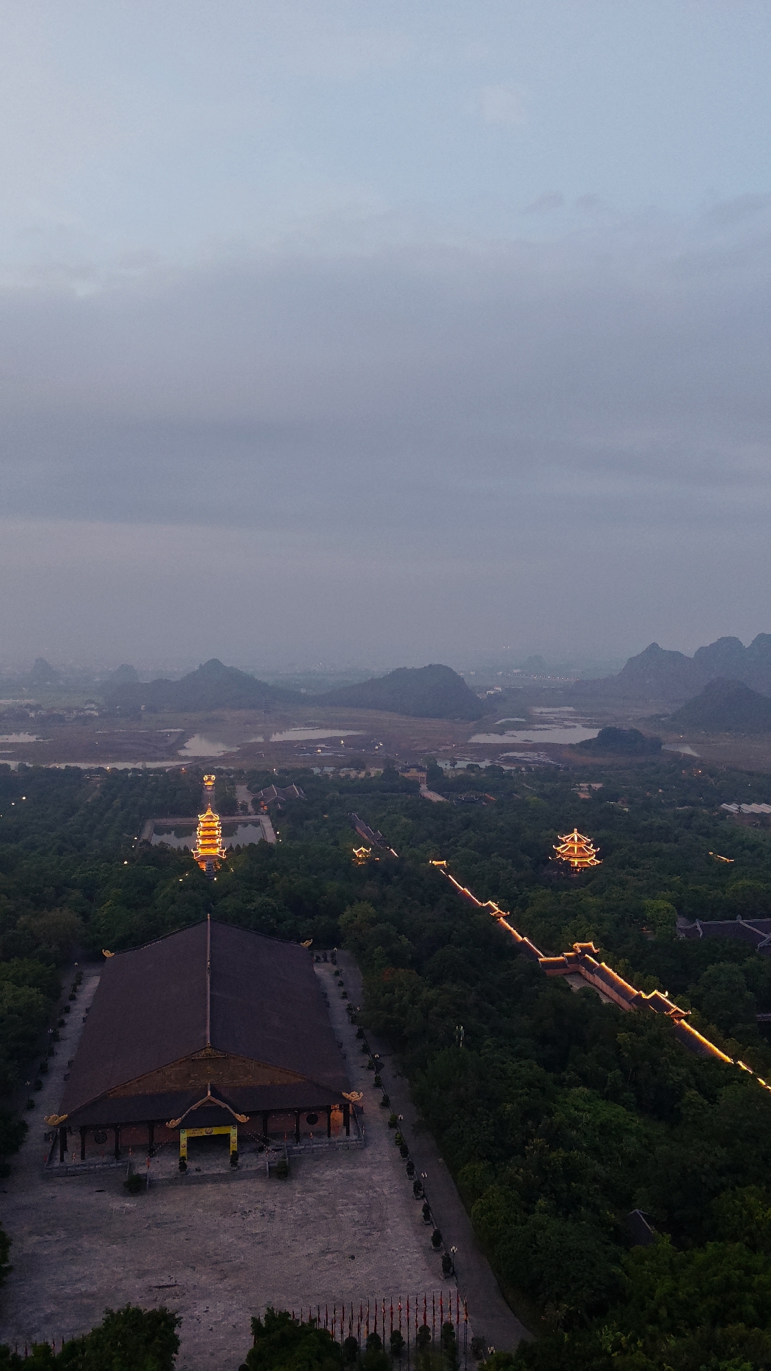 This aerial photograph depicts a twilight scene in the city of Tây Hoa Lư Ward, Vietnam, showcasing a complex of religious and architectural structures nestled within a lush, green landscape. The sky is a soft gradient of pale blue and grey, indicative of dusk or dawn, with diffused clouds.
In the foreground, a large, dark-tiled building with a distinctive, multi-tiered roof dominates the lower left quadrant. Its facade features an entrance marked by yellow banners. The building is surrounded by a large, paved courtyard, sparsely populated by small trees or decorative plants along its perimeter. Rows of flags are visible along the bottom edge of the courtyard.
Further into the mid-ground, the landscape transitions into a dense forest. Within this verdant expanse, several illuminated structures stand out. A prominent, multi-story pagoda, glowing with warm, yellow light, is situated near a body of water. Another smaller, similarly illuminated pagoda is visible to the right of the larger one. A pathway, lined with golden lights, snakes through the trees, connecting different parts of the complex and accentuating the terrain.
In the background, a hazy panorama of karst mountains rises against the muted sky. Ponds and bodies of water are scattered amongst the foothills, reflecting the dim light. The distant horizon suggests a sprawling urban or rural area, softened by the atmospheric conditions. The overall impression is one of serene grandeur and architectural beauty integrated with the natural environment.