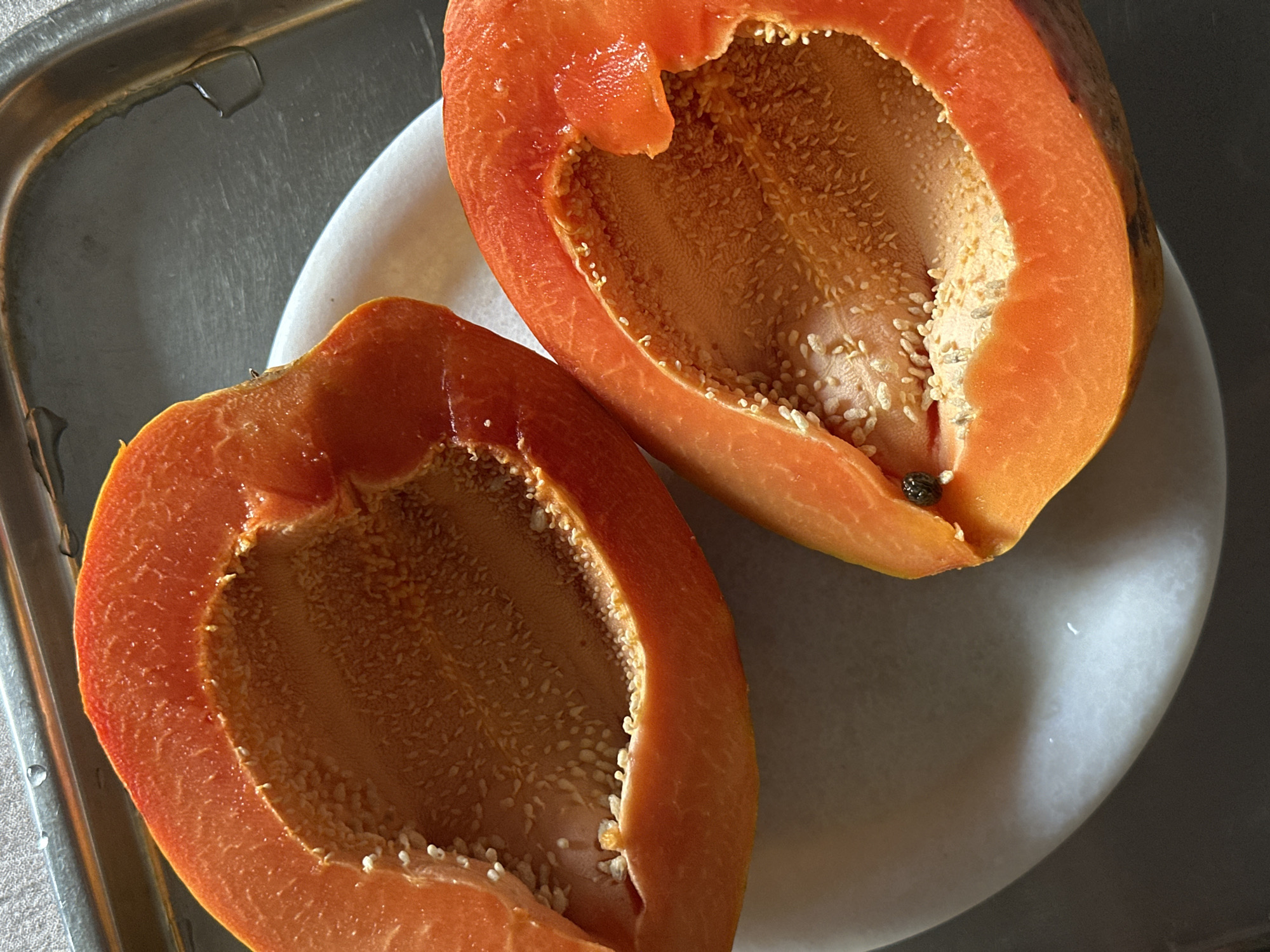 The image depicts two halves of a cut papaya fruit. The papaya is orange and filled with numerous white seeds. The fruit halves are resting on a white plate, which is placed on a silver tray. The lighting suggests an indoor setting. There is no evidence to connect this image to Dharamshala, India.