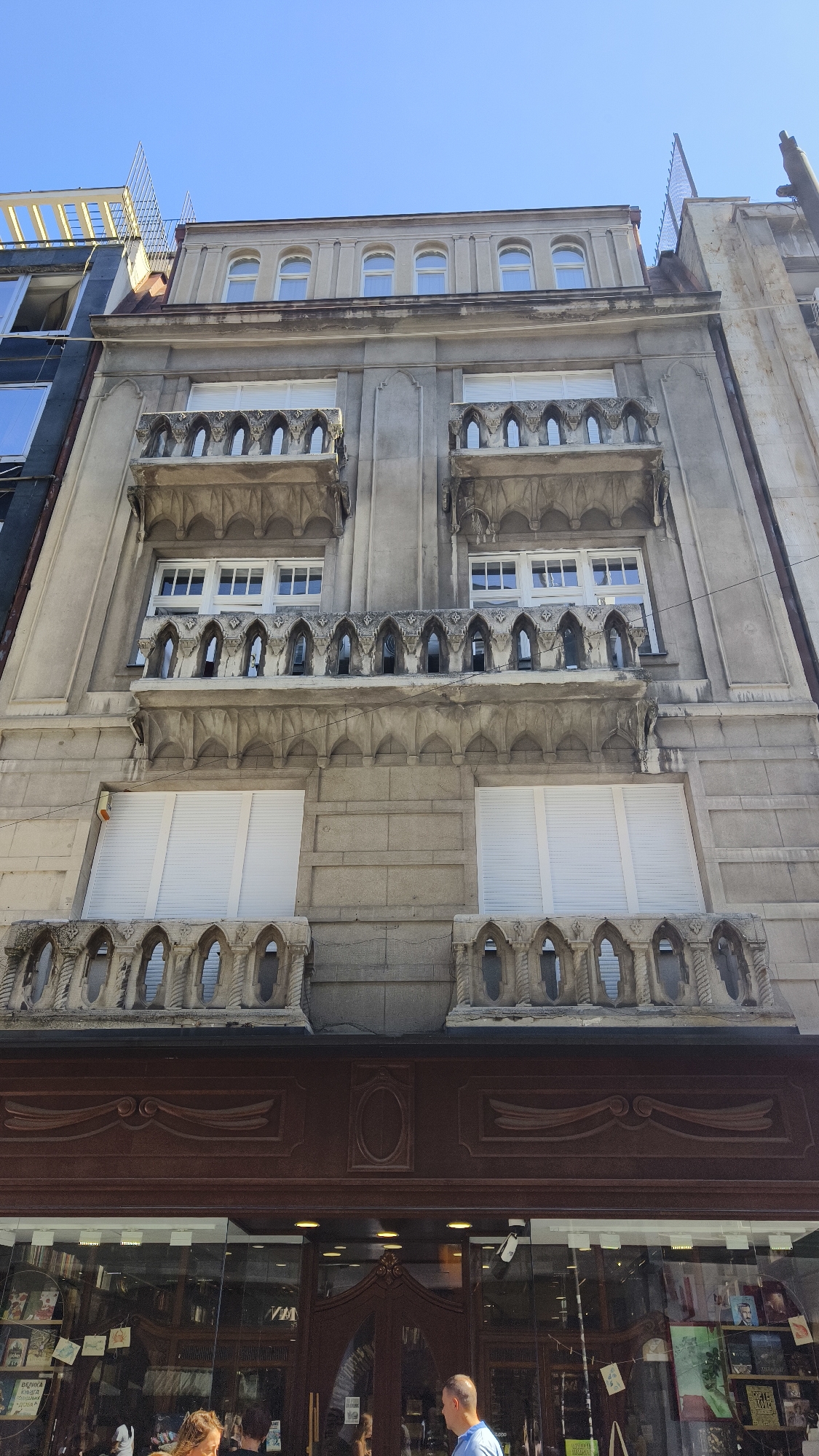 A low-angle, wide shot captures the ornate facade of a stone building with multiple levels of balconies, windows, and decorative stonework. The sky above is a clear, bright blue. The building's upper floors feature arched windows and decorative balconies with intricate carvings. Below these, two rows of large, white-shuttered windows are visible. The lowest visible section of the building is the top of a storefront with large glass windows displaying books and other merchandise. A dark brown, carved wooden fascia runs across the storefront. In the foreground, two individuals are partially visible: a woman with blonde hair on the left and a man in a blue shirt on the right, both appearing to be patrons of the store. The scene is set in an urban environment.