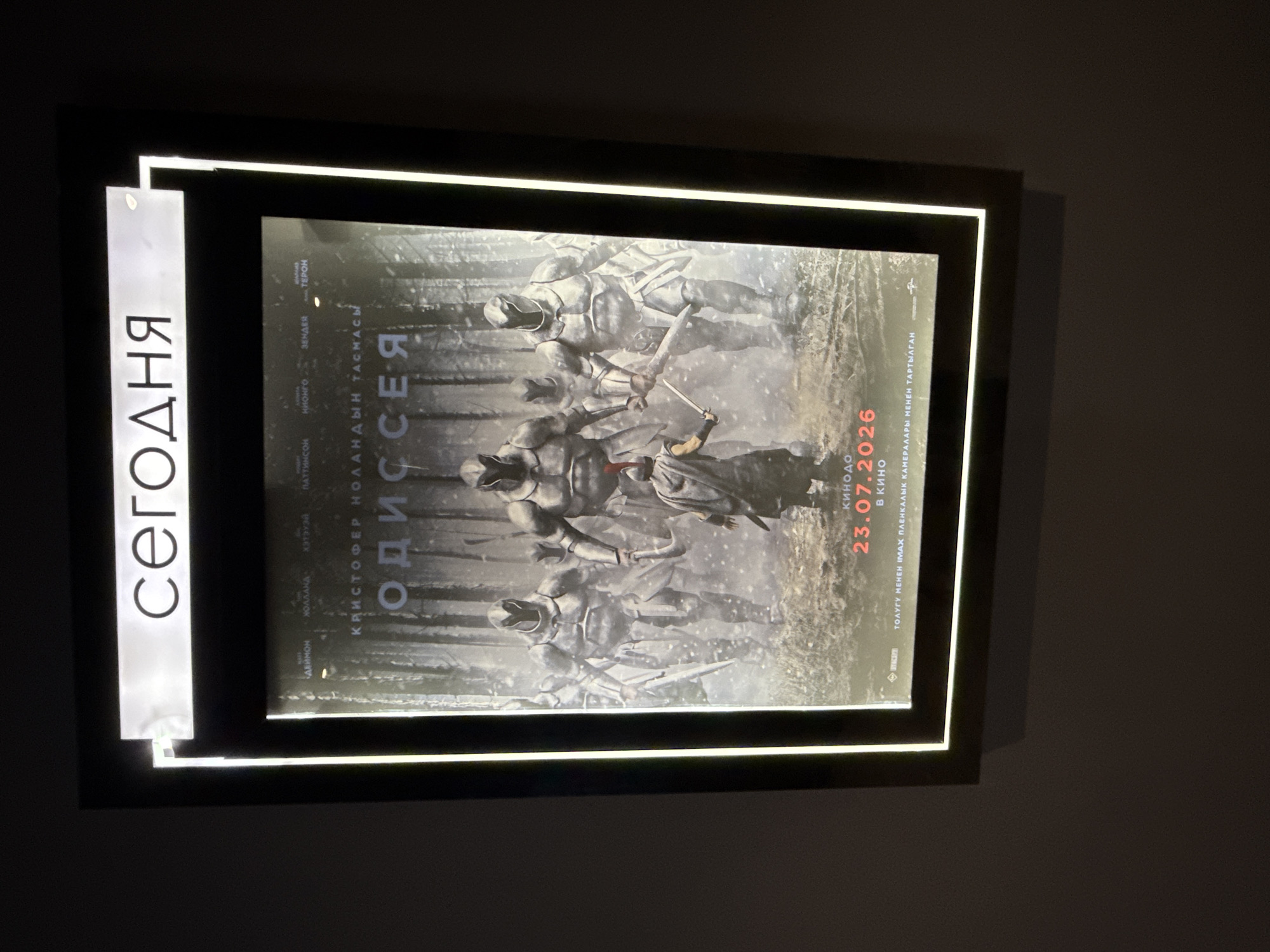 The image shows a movie poster displayed in a backlit frame, likely in a cinema lobby in Bishkek, Kyrgyzstan. The poster features the title "ОДИССЕЯ" (Odyssey) in large, stylized font. Below the title, the names "КРИСТОФЕР НОЛАНДЫН ТАСМАСЫ" (Christopher Nolan's Film) are visible. The artwork depicts a group of figures in metallic armor, resembling soldiers or warriors, in a misty, wooded environment. In the foreground, a smaller figure in a helmet and cape, holding a sword, stands facing the armored figures. The release date "23.07.2026" is prominently displayed in red text, along with "В КИНО" (In Cinema). At the bottom of the poster, there is text in what appears to be Kazakh or a similar Turkic language, reading "ТОЛУҒЫ МЕНЕН IMAX КАМЕРАЛАРЫ МЕНЕН ТАРТЫЛҒАН" which translates to "Filmed entirely with IMAX cameras". The overall presentation suggests a promotional display for an upcoming film.