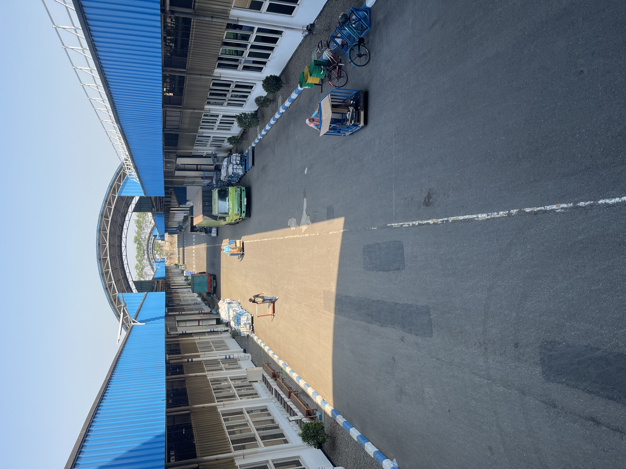 The image depicts an outdoor scene at what appears to be a logistics or manufacturing facility in Pelang, Indonesia. The perspective is from an elevated vantage point, looking down on a paved roadway flanked by industrial buildings. The buildings feature blue corrugated metal roofs and siding, with white window frames. Along the left side of the roadway, there are rows of wooden benches and a small tree. The roadway itself is marked with white lines.
Several individuals and objects are present. On the left side of the roadway, a person is pushing a wheeled cart loaded with white sacks. Further down the road, a green truck is parked. On the right side of the roadway, a person wearing a pink headscarf is inside a blue cage-like structure on wheels, possibly being moved or transported. Two bicycles are parked near the cage structure, and there are green and yellow trash bins. The sky is clear and blue, indicating a sunny day. The overall environment suggests a functional industrial area with ongoing activity.