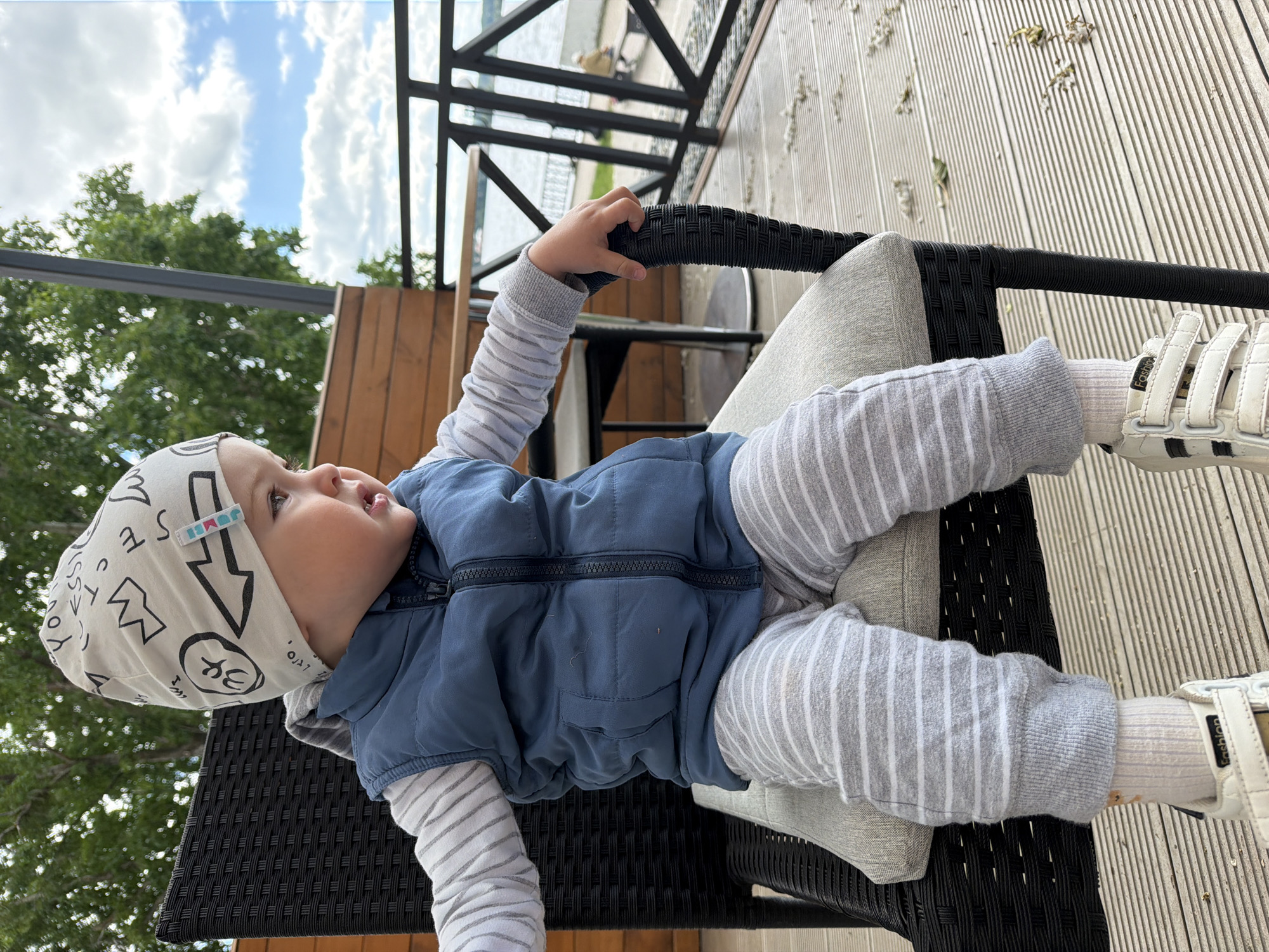 A young child, likely a toddler, is seated on a black woven chair on a wooden deck. The child is wearing a white beanie with a black graphic print, a blue puffer vest over a striped long-sleeved shirt, grey striped pants, white socks, and white sneakers. The child is looking to the right, with their right hand resting on the armrest of the chair.
In the background, there is a railing and a body of water, possibly a lake or river. Trees are visible on the left, and a cloudy sky is present. The overall setting appears to be an outdoor seating area, likely a cafe or restaurant with a view of the water. The scene is well-lit, suggesting it is daytime.
The location is identified as Votkinsk, Russia.
