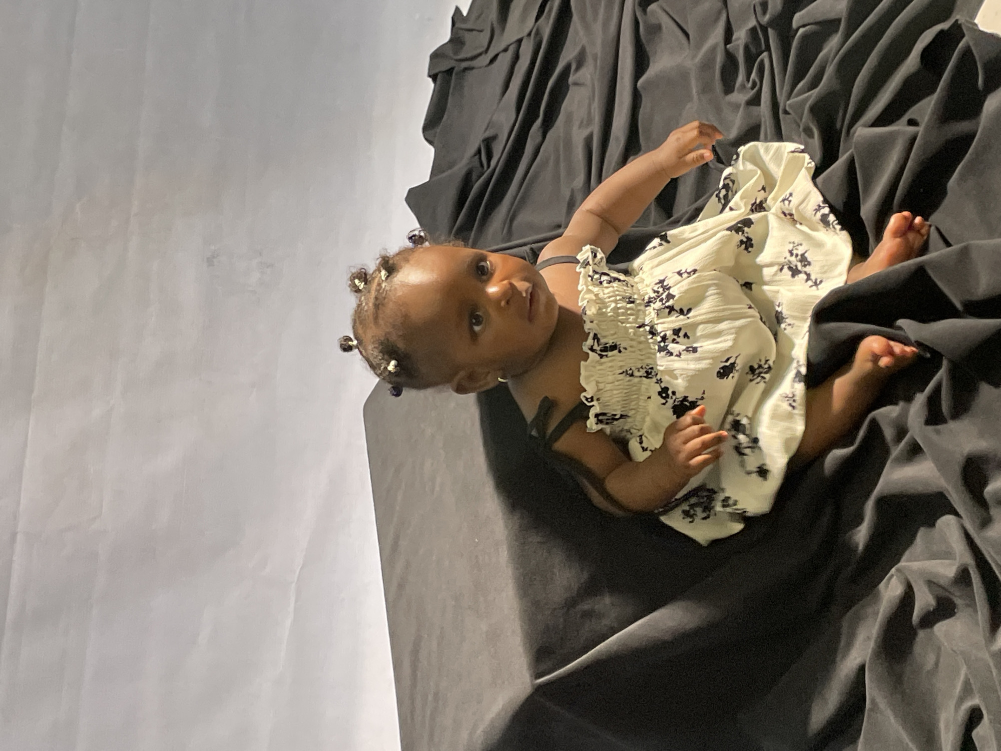 A baby lies on her side amidst draped black fabric. She wears a white dress with a black floral pattern and has small, decorative hair ties. She is facing the camera. The backdrop is a light gray. The location is unknown and cannot be determined to be Jos, Nigeria from the image.