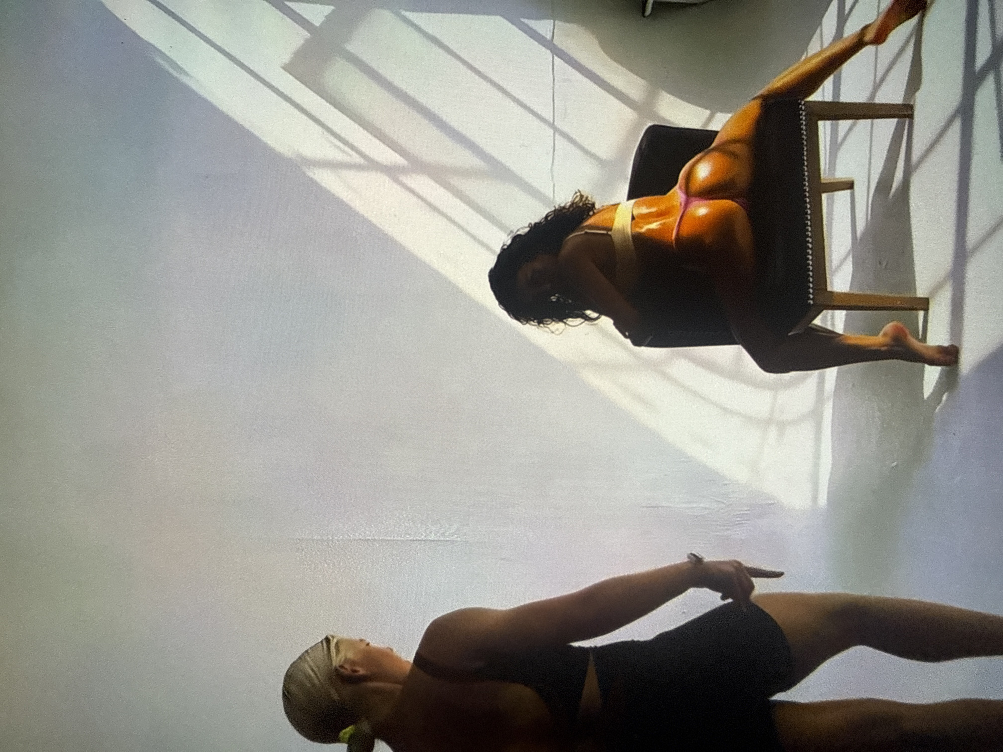 The image depicts an indoor scene within a minimalist studio environment located in Varna, Bulgaria. Two individuals, both women, are present. On the left, a woman with blonde hair tied back, wearing a dark sports bra and dark shorts, stands with her back mostly to the viewer. Her head is turned slightly to the right, suggesting she is observing the other individual. Her right arm is bent at the elbow, with her hand resting near her thigh. On the right side of the frame, a second woman with dark, curly hair is seated on a dark, upholstered chair. She wears a light-colored bralette and bright pink thong-style bottoms. Her body is angled away from the viewer, but she has turned her head over her left shoulder, looking towards the camera. Her left leg is bent, with her foot near the chair, while her right leg is extended outwards to the right. The background consists of plain, light-colored walls and a matching floor, typical of a photography studio. Strong, directional light, likely natural sunlight entering from an off-camera window to the right, casts distinct, grid-like shadows across the right wall and floor, creating dramatic contrasts and highlighting the seated woman's form.
