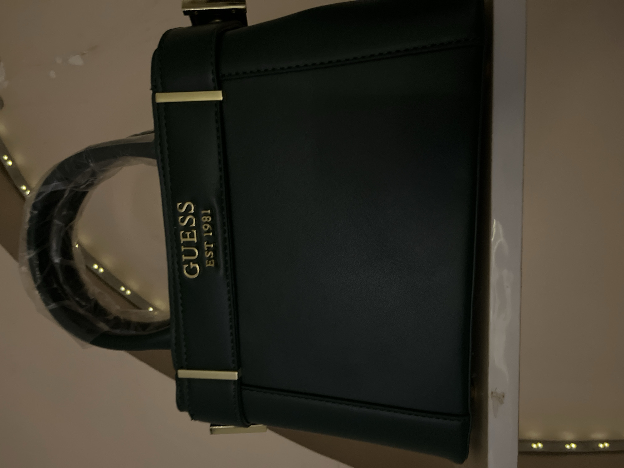 A dark green Guess handbag with gold-toned hardware and the "GUESS EST 1981" logo is displayed on a white shelf. The handbag has a structured shape with top handles and a flap closure. The background is a light beige wall, and the scene is illuminated by string lights, creating a warm ambiance. The image was taken indoors, likely in a residential setting. The location is not specified within the city of Abuja, Nigeria.