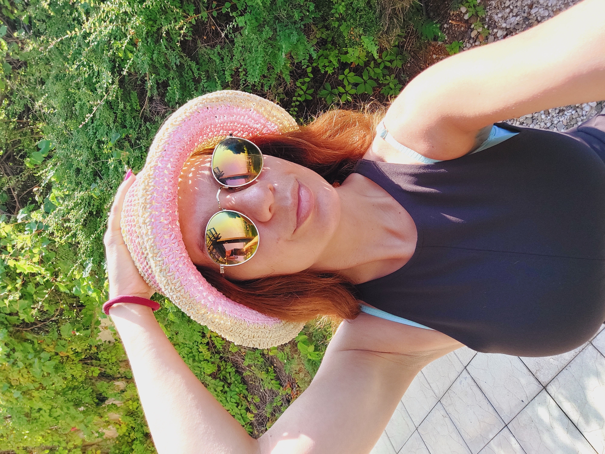 A woman is taking a selfie outdoors. She is wearing a straw hat with pink and yellow accents, sunglasses, and a dark tank top. Her right arm is raised, holding the hat. The background consists of lush green foliage and a paved walkway. The reflection in her sunglasses shows a treed area and a body of water, possibly a harbor, consistent with the coastal city of Sozopol, Bulgaria.