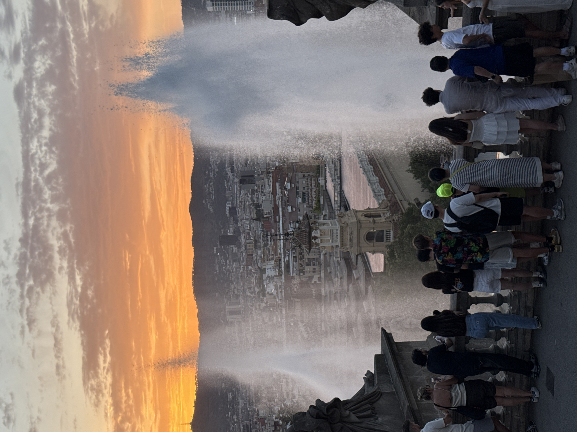 The image shows a group of people standing on a raised platform, overlooking a cityscape with a large fountain. The sun is setting, casting an orange glow on the sky and silhouetting the hills in the background. The fountain is spraying water high into the air, creating a dramatic effect. The city appears to be Barcelona, Spain, based on the architecture and the distinctive topography. The people in the foreground are diverse in age and dress, with some holding phones and cameras, suggesting they are tourists or locals enjoying the view. The overall scene is one of awe and appreciation for the natural and man-made beauty of the city.