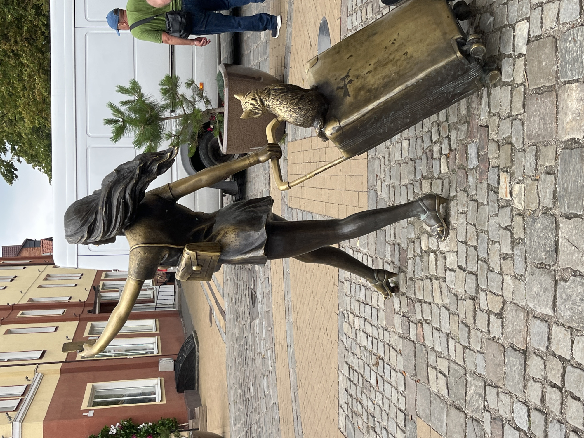 The image depicts a bronze sculpture of a woman taking a selfie with a cat perched on her suitcase. The sculpture is located in Zelenogradsk, Russia, on a cobblestone street. The woman is depicted in a dynamic pose, with her arm extended to take a picture. She is wearing a dress and high heels. The cat is sitting on top of the suitcase, looking away from the camera. In the background, a white van and a building with a red and yellow facade are visible. A man in a green t-shirt and blue jeans is walking away from the camera in the background. The environment appears to be an urban setting, with trees and potted plants adding to the scenery.