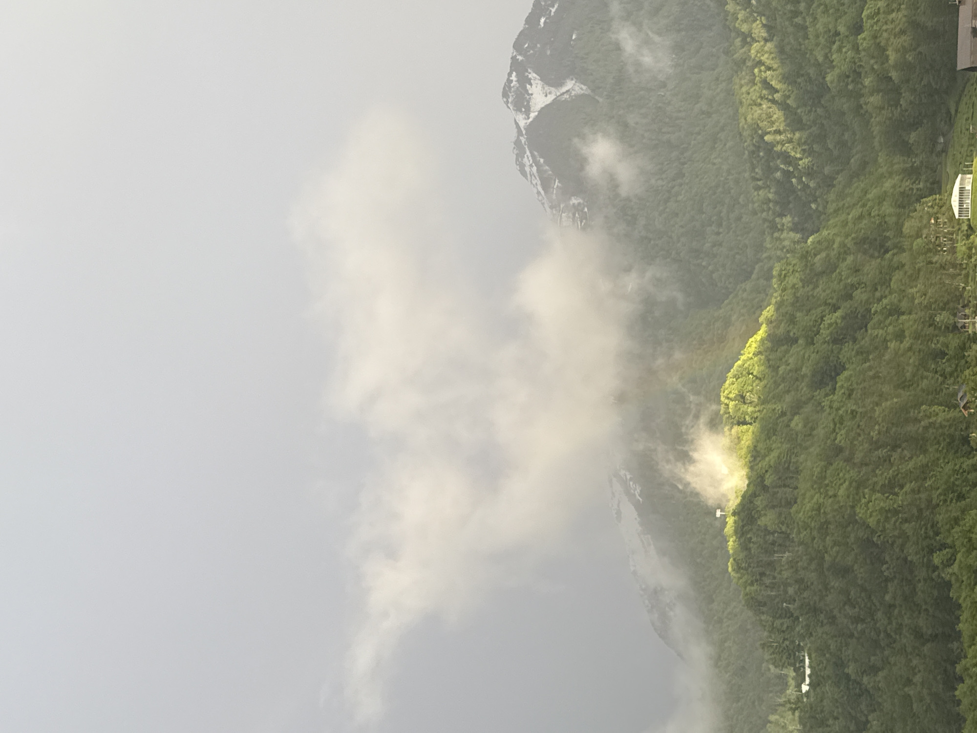 The image shows a mountainous landscape in Krasnaya Polyana, Russia, partly obscured by clouds. A prominent mountain peak is visible on the right, with snow near its summit. Below the peak, dense green vegetation covers the mountain slopes. Light penetrates through gaps in the clouds, illuminating parts of the forest. A small structure, possibly a building or observation point, is visible on the slope amongst the trees. The left side of the image is dominated by cloudy skies.