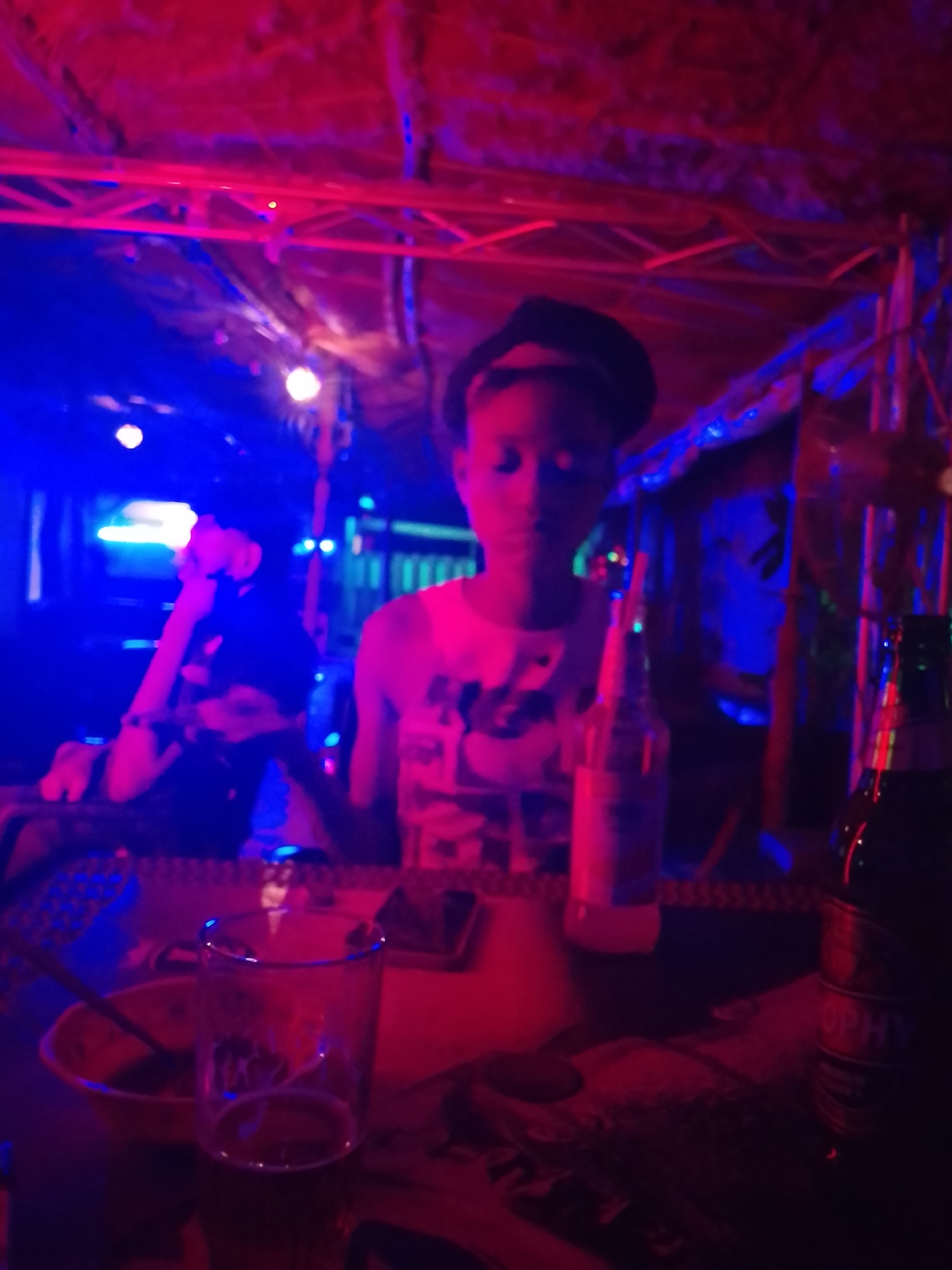 An indoor photograph captures a scene illuminated by strong artificial blue and red lighting, suggesting a nighttime setting such as a bar, club, or casual venue.
In the foreground, a person is seated at a table, facing slightly towards the camera. They wear a dark cap or hat and a light-colored top. Their face is partially obscured by shadows and the colored lighting. To their left, a second person is visible in the blurred background, also seated and seemingly looking to their side.
On the table in front of the primary subject, several objects are visible. There is a clear drinking glass, holding a small amount of liquid at the bottom. Next to it, a small orange bowl contains a dark substance and a spoon or utensil. Further to the right, a clear plastic bottle stands, and beside it, a dark glass bottle with a label partially visible, showing the letters "OPHY" (likely part of a brand name like "Trophy"). A smartphone might also be present on the table, partially hidden.
The background features metal trussing or scaffolding-like structures supporting what appears to be a rustic or makeshift ceiling, possibly made of woven material or corrugated metal, heavily bathed in red light. Bright blue and red lights are scattered throughout the space, casting vibrant hues on the subjects and surroundings. The overall atmosphere is relaxed and social, typical of an evening gathering spot.