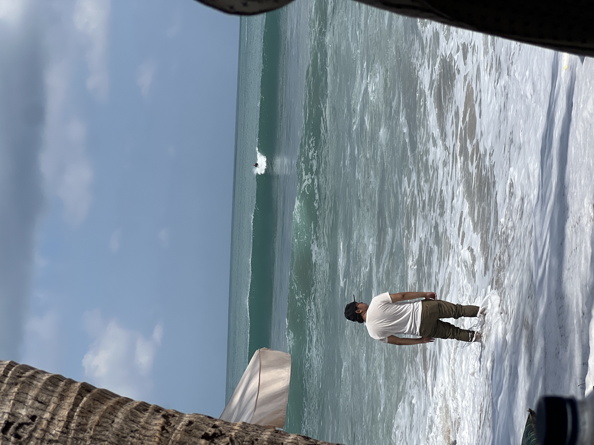 A man stands in shallow ocean water at a beach in Kuta, Indonesia, facing away from the camera. He wears a white t-shirt and olive green pants. The ocean has turquoise waves with white foam in the foreground and deeper green water beyond. A surfer is visible on a wave in the distance. A large palm tree trunk is on the left side of the image, and a portion of a light-colored umbrella is visible behind it. The sky is blue with scattered white clouds.