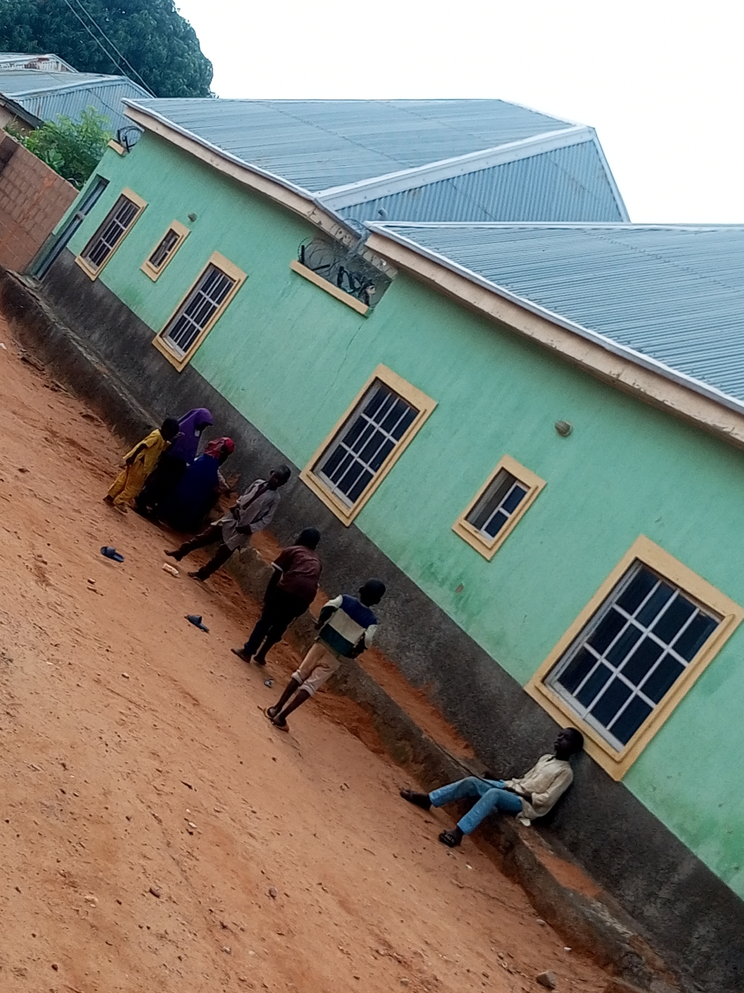 The image depicts a residential area in Tempure, Nigeria. The foreground is dominated by a sloping dirt path, with scattered stones and a few dark-colored flip-flops. In the midground, a group of individuals are present. On the left, two children, one in a yellow outfit and another in purple and dark blue clothing, are near a wall. A man in a grey tunic and trousers stands in the center, facing away from the viewer, with his arms bent. To his right, another individual in a burgundy top and dark trousers is also facing away. Further to the right, a person in a striped long-sleeved shirt and light-colored shorts stands. In the far right of the midground, a young man in a light-colored shirt and blue jeans is seated, leaning against the wall of a building. The background features a row of light green buildings with corrugated metal roofs. The buildings have several windows with yellow frames. The overall scene suggests a casual, outdoor gathering in a community setting.