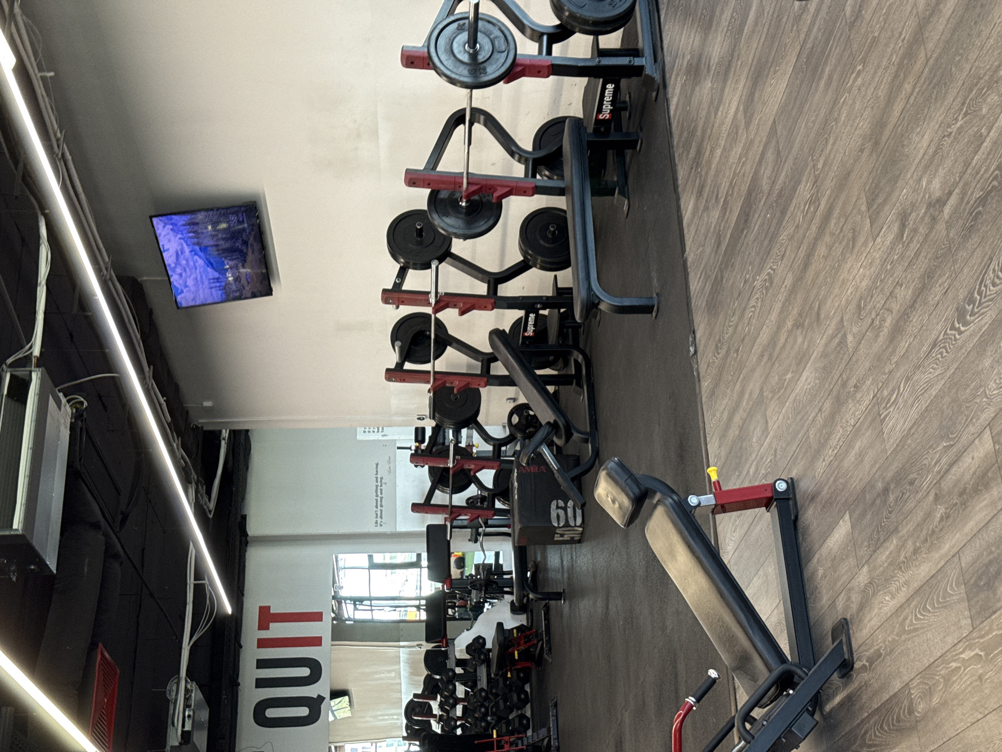 The image shows the interior of a gym in Sofia, Bulgaria. Weightlifting equipment is prominently displayed, including weight racks and benches. A television is mounted on the ceiling. The word "QUIT" is visible on a wall sign. The floor is covered with a wood-grain-patterned material. The overall environment appears to be well-lit and modern. No people are visible in the frame.