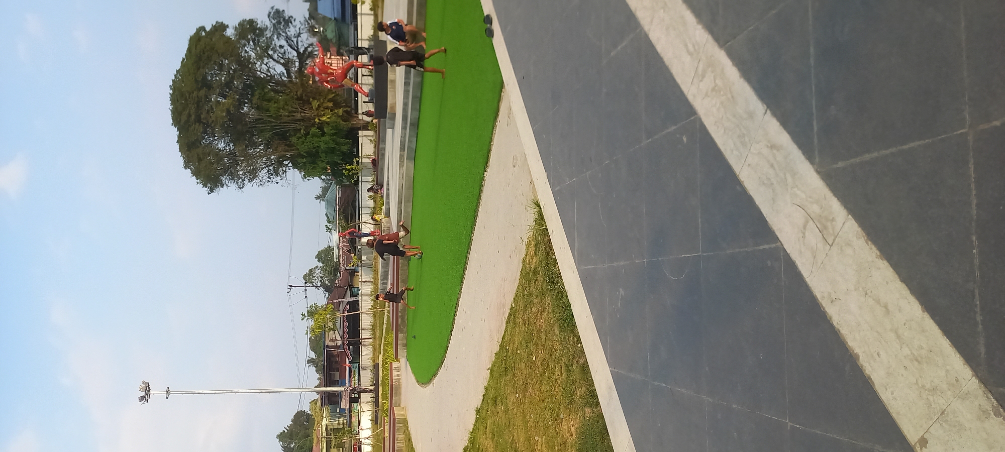 This image depicts an outdoor public space in Melayu, Indonesia, characterized by a mix of recreational areas and urban elements. The foreground features a dark gray tiled plaza with a light-colored inlaid strip, suggesting a modern design. Beyond this, a curved pathway leads to a vibrant green artificial turf area. On the turf, several young people are engaged in playing soccer, with one boy in the process of kicking the ball.
In the background, a prominent statue of Iron Man stands on a raised platform, flanked by a smaller statue of Spider-Man. The park is bordered by a low wall and a light pole is visible on the left side. Further in the distance, a row of buildings forms the urban backdrop, with a large, leafy tree dominating the right side of the frame. The sky is a clear light blue with a few wispy clouds, indicating daytime. The overall scene suggests a community gathering space where leisure activities take place amidst an urban environment.