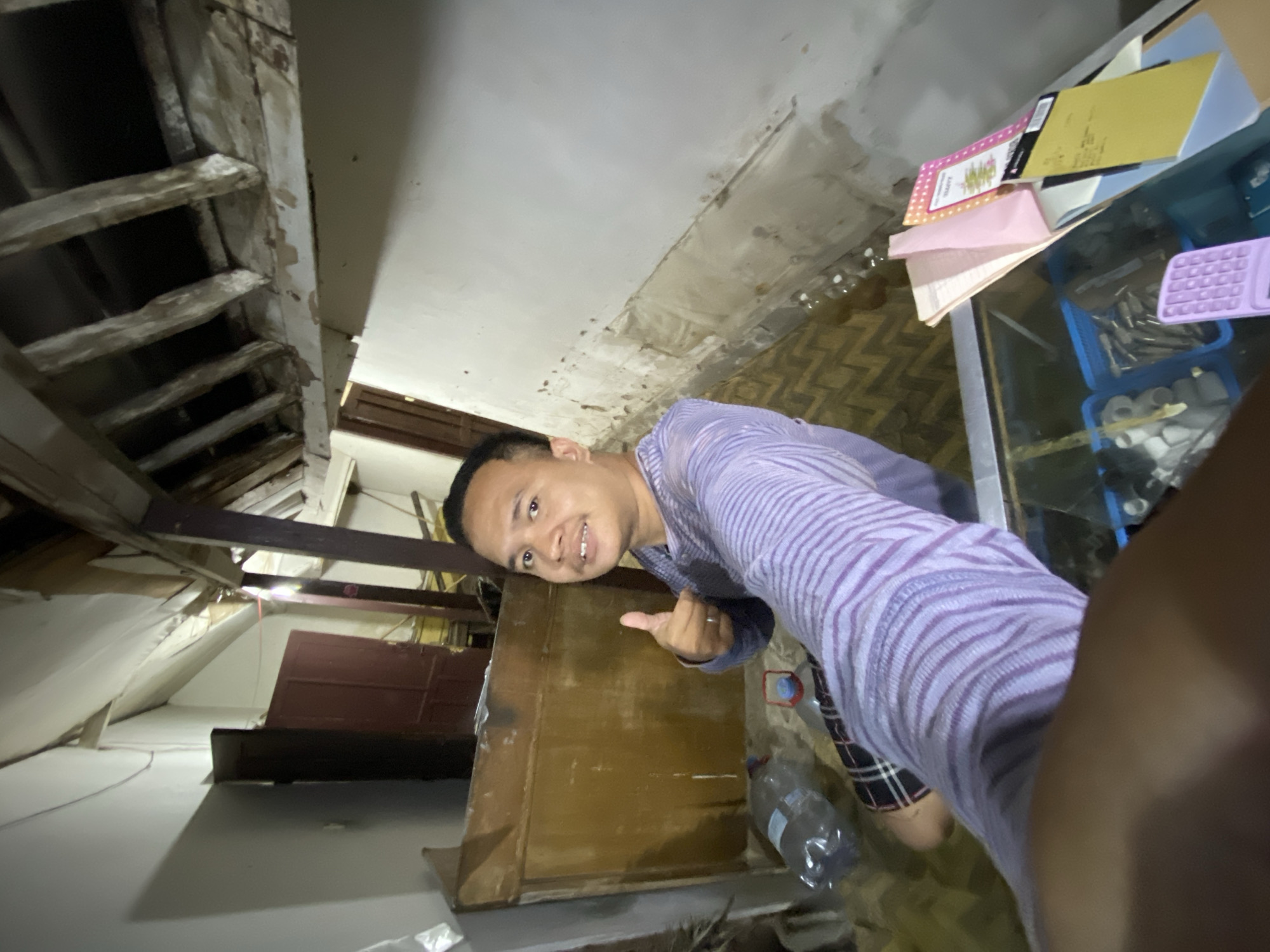 In the city of Sugihwaras, Indonesia, a man is captured in a selfie taken from a low angle, holding his right thumb up in a gesture of approval. He is wearing a long-sleeved purple shirt with thin horizontal stripes and checkered shorts. The man is positioned in what appears to be an unfinished or dilapidated room. To his left, a large wooden structure, possibly a cabinet or partition, stands against a white wall. In front of the wooden structure, several plastic water bottles are scattered on a woven mat. The ceiling of the room is unfinished, with exposed wooden beams and what appears to be a white cloth stretched across some of them. To the right of the man, a glass-topped display case holds various items, including papers, a blue plastic container filled with small objects, and a purple calculator-like device. The overall environment suggests a construction site, storage area, or a poorly maintained living space.
