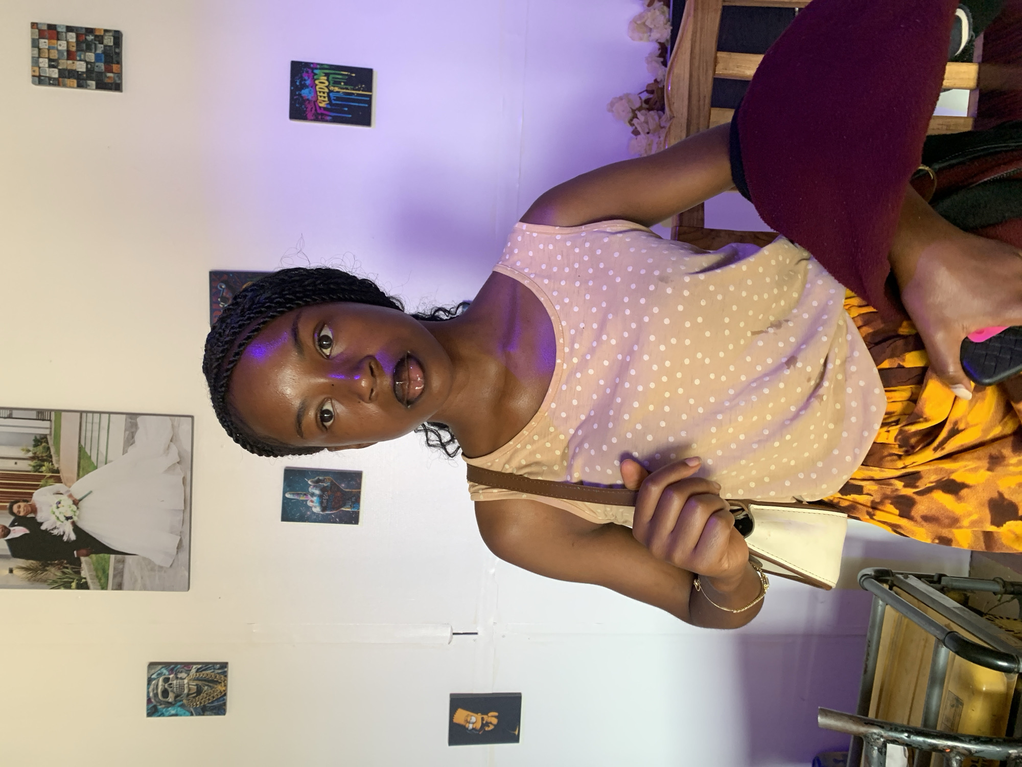 A young Black woman is captured in a close-up, rotated portrait, seemingly indoors in Jos, Nigeria. She is looking directly at the camera with her mouth slightly open, suggesting she might be mid-conversation or posing. Her hair is styled in braids. She wears a sleeveless, light peach or beige top adorned with white polka dots, and a patterned skirt with brown and yellow/orange hues, possibly an animal print. A dark red fabric, possibly a blanket or garment, rests on her lap. She carries a light-colored shoulder bag with a brown strap, which she holds with her right hand. On her left wrist, she wears a delicate gold-colored chain bracelet, and she holds a black phone with a pink accent in her left hand.
The background is a white wall decorated with several small pictures or canvases. Notable artworks include a mosaic-like pattern, a dark piece with "FREEDOM" written in colorful, dripping letters, and a larger vertical picture depicting a wedding couple—a bride in a white gown with a bouquet and a groom in a dark suit. Other smaller pictures show a skull with a gold chain, a stylized hand with colorful elements, and a cartoon character (possibly Bart Simpson). The upper part of the wall exhibits a distinct purple/violet glow, indicative of ambient mood lighting, perhaps from LED strips. Parts of a wooden furniture piece, possibly a chair or bed frame, and white decorative flowers are visible behind her. To the far right, a metal-framed object, possibly a table or stand, is partially seen. The setting appears to be a personal room or a casual indoor space.