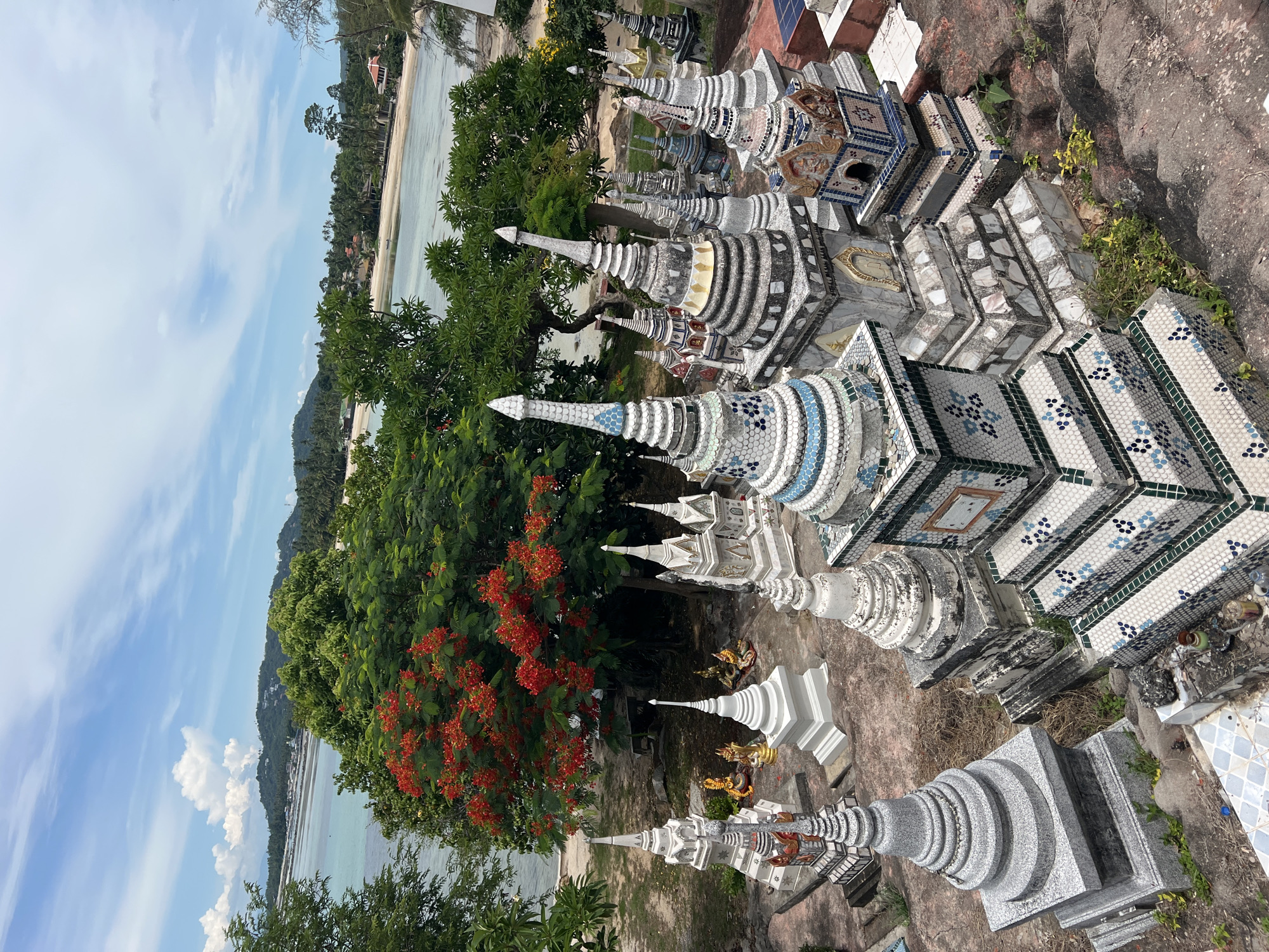 A high-angle, wide shot shows a collection of tiered, decorative stupas arranged on a dusty, uneven ground. The stupas are constructed from concrete and adorned with mosaic tiles in white, blue, and green patterns. Some stupas have small golden Buddha statues placed around them. In the background, a lush green tree with vibrant red flowers stands out against a bright blue sky with scattered white clouds. Beyond the tree, a body of water, likely the sea, is visible, with a distant coastline dotted with buildings and palm trees. The scene appears to be located outdoors in a tropical environment, possibly a temple or memorial site in Baan Hua Thanon, Thailand. No people are present in the image.