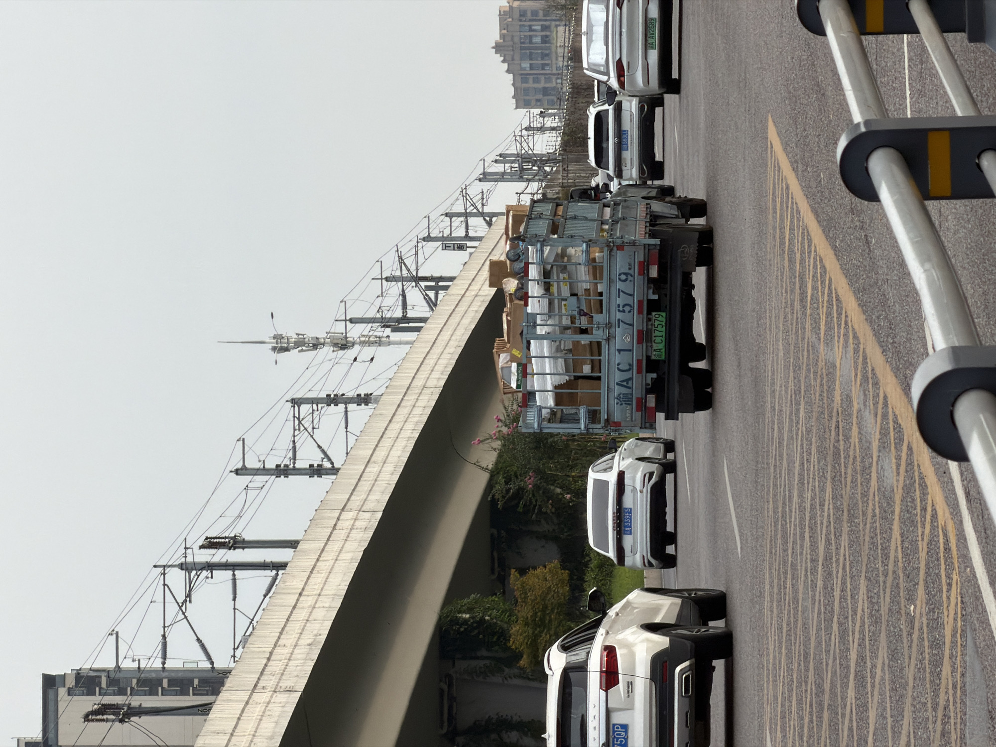 This image captures a street scene in Jiangjin, China, under an overpass supporting railway tracks. Several vehicles are visible on the road, including a white SUV on the left, a white sedan in the middle, a light blue truck carrying boxes, and another white sedan on the right. Overhead power lines for the railway stretch across the frame, supported by poles. The road surface features yellow and white lane markings. Buildings are visible in the background, and the sky is overcast. The overall impression is of a busy urban environment.