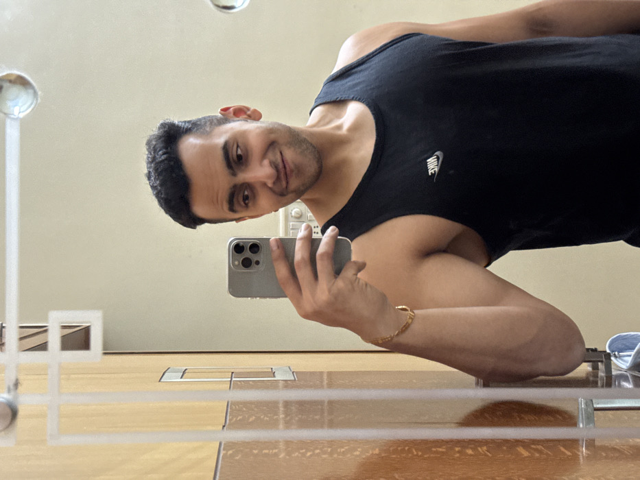A man takes a mirror selfie. He is muscular and wearing a black tank top with a Nike logo. He holds a grey iPhone with three camera lenses. A gold bracelet adorns his wrist. The reflection reveals a tiled floor, an electrical outlet, and a glass shelf or frame. The man gazes directly at the camera with a slight smile.