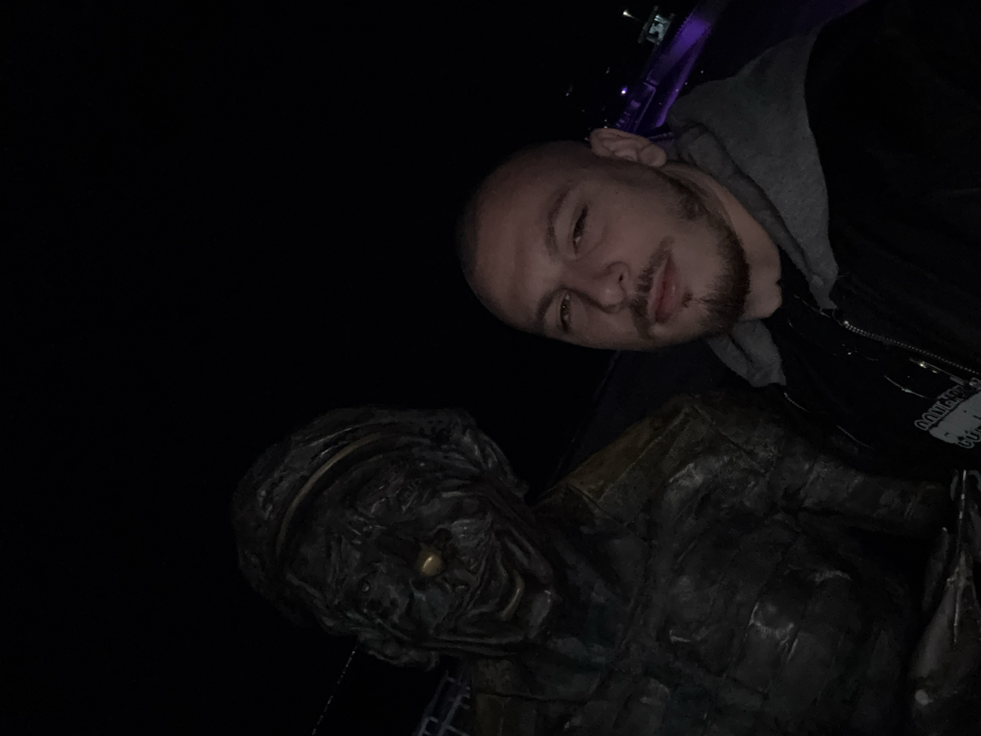 A close-up, slightly angled photograph shows a man next to a bronze sculpture. The man has a shaved head, light skin, and a short beard. He is wearing a hooded sweatshirt with a black jacket. The sculpture appears to be a depiction of an older person wearing a cap or hat. The background is dark, suggesting it is nighttime. The sculpture may be of Alyosha, a character in a story by famous Bulgarian writer Georgi Baev. It is located at the Sea Garden in Burgas, Bulgaria.