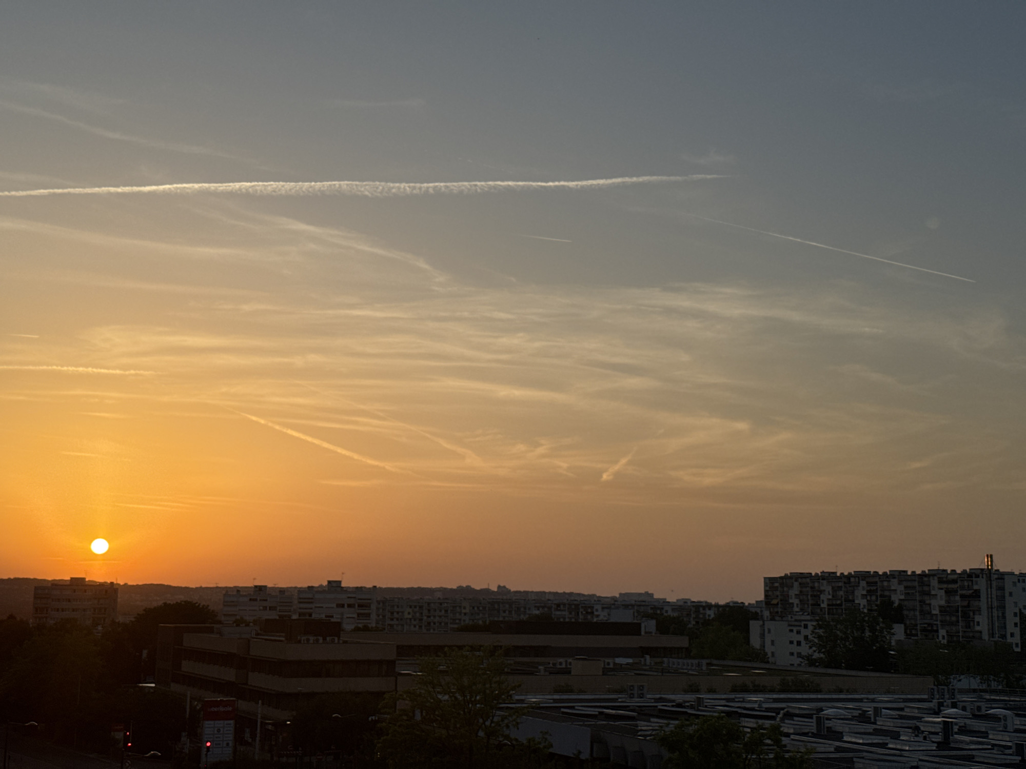 The image depicts an urban skyline at sunset. The sun, a bright orange disc, is low on the horizon, casting a warm, golden glow across the sky. Wispy clouds, tinged with orange and pink, are scattered throughout the sky, with prominent contrails from airplanes streaking across the atmosphere. In the foreground, silhouetted buildings of varying heights create a dense urban landscape. Some buildings appear to be residential blocks with multiple windows, while others seem to be commercial or industrial structures with flat roofs. Trees, also in silhouette, are interspersed among the buildings. The overall impression is one of a city at dusk, bathed in the soft light of the setting sun.