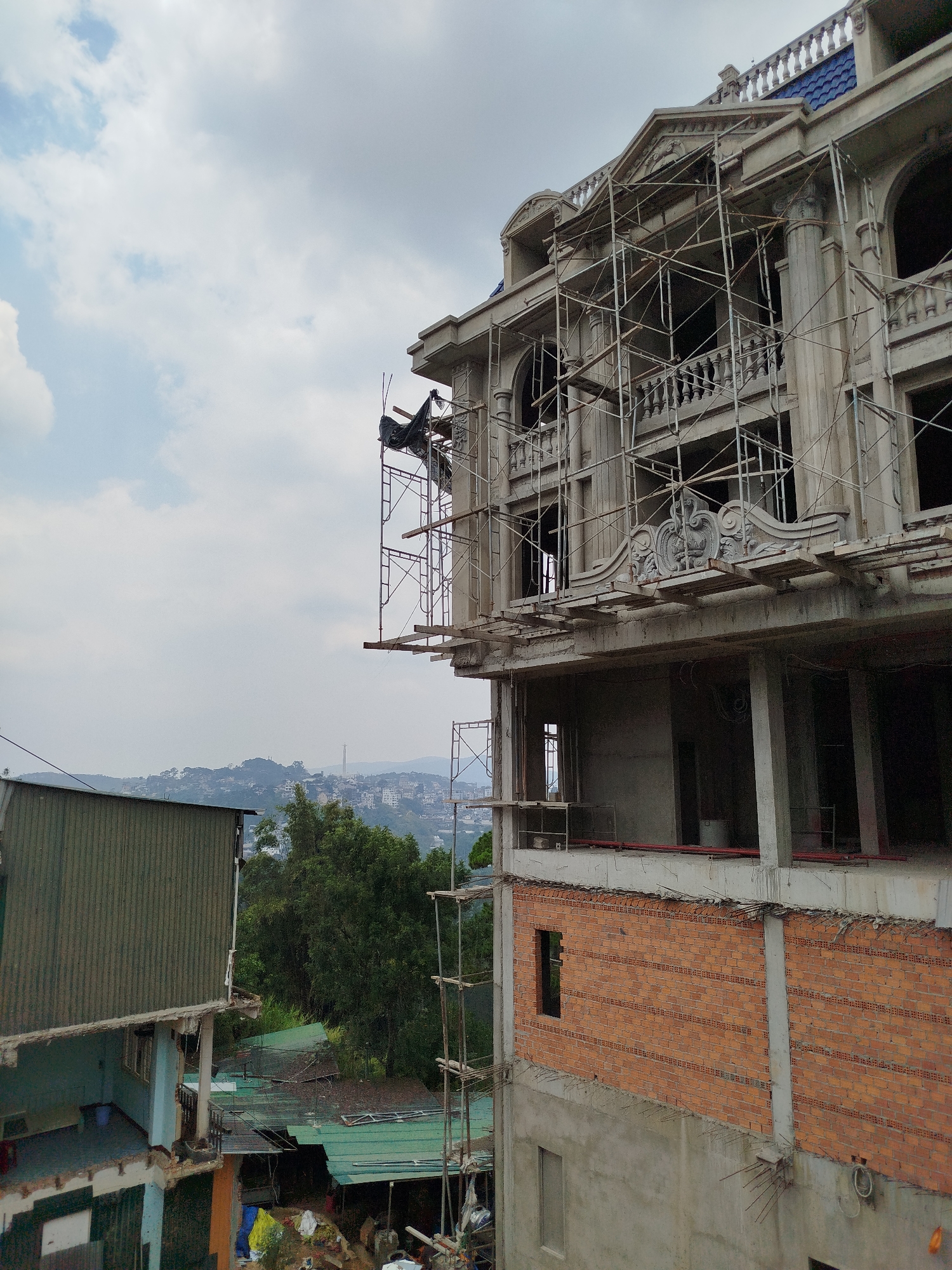 The image depicts a building under construction in Da Lat, Vietnam. The structure is multi-storied, with the lower levels showing brick and concrete construction. The upper level features ornate decorative elements and is surrounded by scaffolding. A smaller, dilapidated building is adjacent to the construction site. In the background, the city of Da Lat is visible with its characteristic hilly terrain. The sky is overcast with a mix of clouds. There are no visible people in the frame.