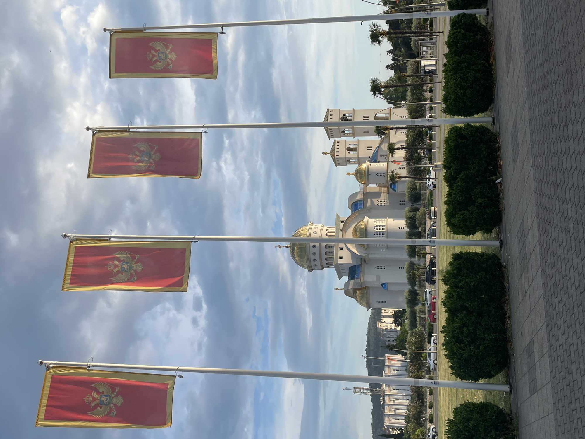The image depicts four Montenegrin flags waving from flagpoles, with the Cathedral of Saint John Vladimir in the background. The flags are red with a gold border and feature the Montenegrin coat of arms. The cathedral is a large, ornate structure with gold domes and white walls. The scene is captured outdoors, showing a paved area, manicured bushes, palm trees, and other elements suggesting a public space. The location is the city of Bar, Montenegro. There are no visible people in the image. The flags are the primary subject, with the cathedral serving as a backdrop.