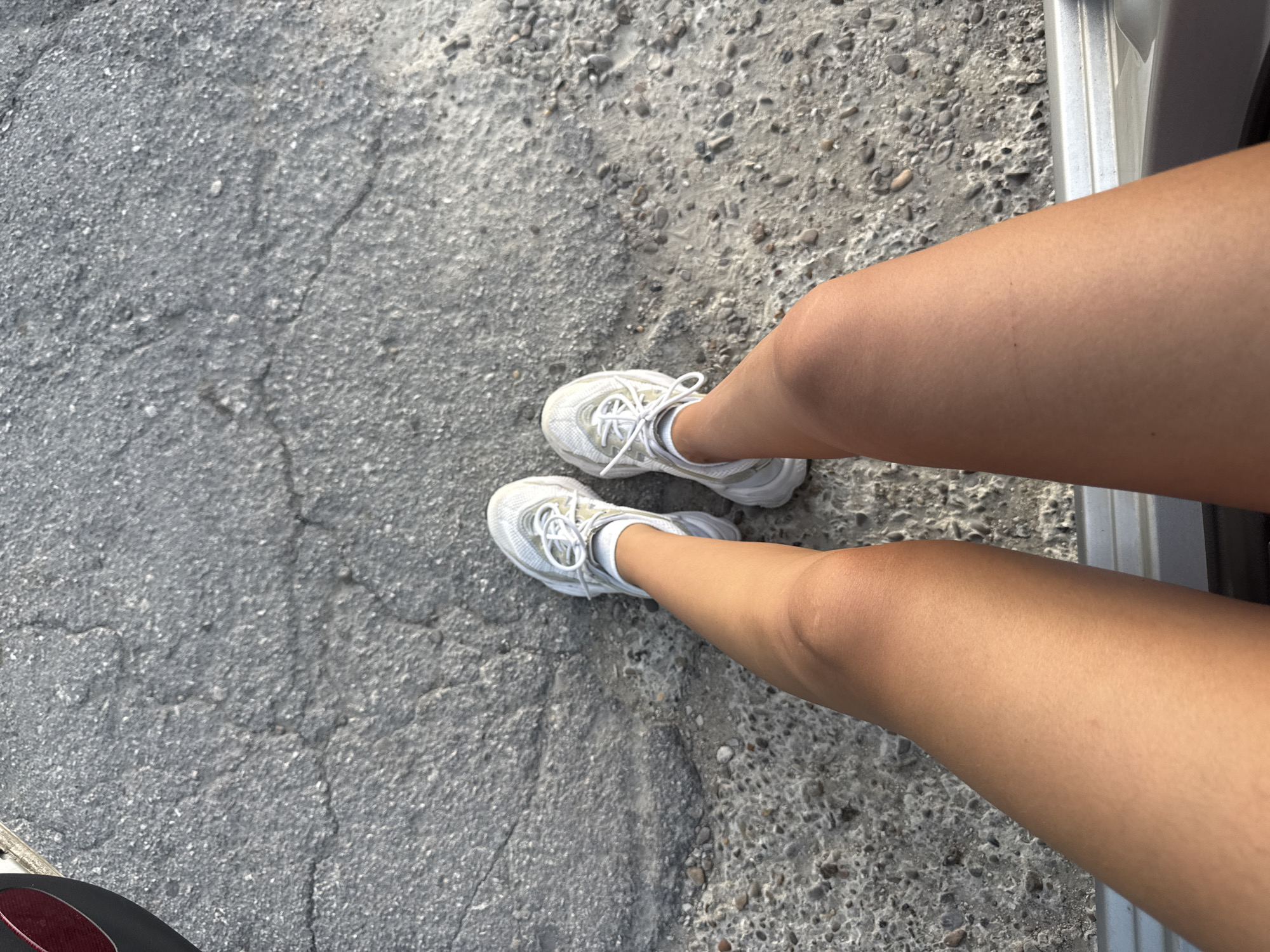 A low-angle, outdoor shot depicts a person's legs from the knees down, standing on a cracked asphalt surface. The person is wearing white sneakers with white laces. The left leg is closer to the camera and the right leg is slightly behind it. The skin tone of the legs is tanned. The environment appears to be urban, with the asphalt possibly being a sidewalk or road. There are small pebbles and debris scattered on the asphalt. The image does not contain enough information to determine the specific location within the city of Cimișlia, Moldova.
