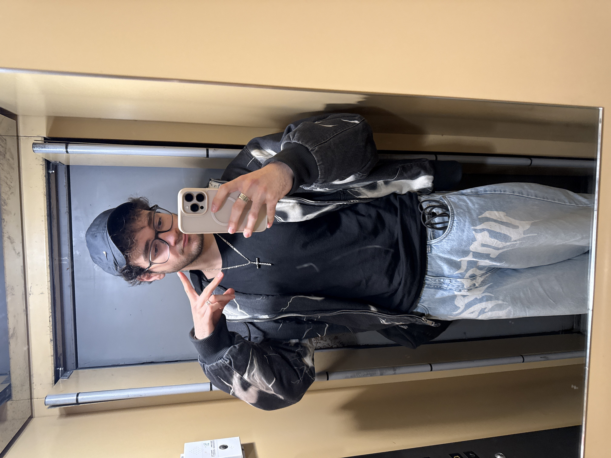 A young adult male is captured in a mirror selfie within an interior space in Blagoevgrad, Bulgaria. He stands facing the mirror, holding a light-colored smartphone with triple camera lenses in his right hand, pointed toward his reflection. His left hand is raised, forming a 'peace' sign with his index and middle fingers extended.
The subject has dark, curly hair and light facial stubble, wearing dark-framed glasses. He is dressed in a dark baseball cap with a light-colored logo (resembling a Nike swoosh), worn backward. Beneath a black crew-neck t-shirt, he wears a dark gray or black zip-up jacket featuring a distinctive white and gray abstract pattern, reminiscent of lightning strikes, across its body and sleeves. The jacket's zipper is partially open. He wears light blue denim jeans with a bleached or distressed appearance, embellished with large white graffiti-style lettering or flame patterns on the front thigh areas.
Accessories include a silver-colored chain necklace with a prominent cross pendant. He wears a textured ring on the ring finger of his left hand and a simple band ring on the ring finger of his right hand. A dark, coiled object, possibly headphones or cables, is visible in the right front pocket of his jeans.
The immediate environment visible in the reflection consists of the mirror in the foreground. In the background, there is a dark gray door, and a slender, silver-colored metallic pole with two black bands runs vertically along the left side of the reflected scene. Walls appear to be a light beige or pale orange hue, and a white wall-mounted panel with buttons or indicators is partially visible on the far left.
