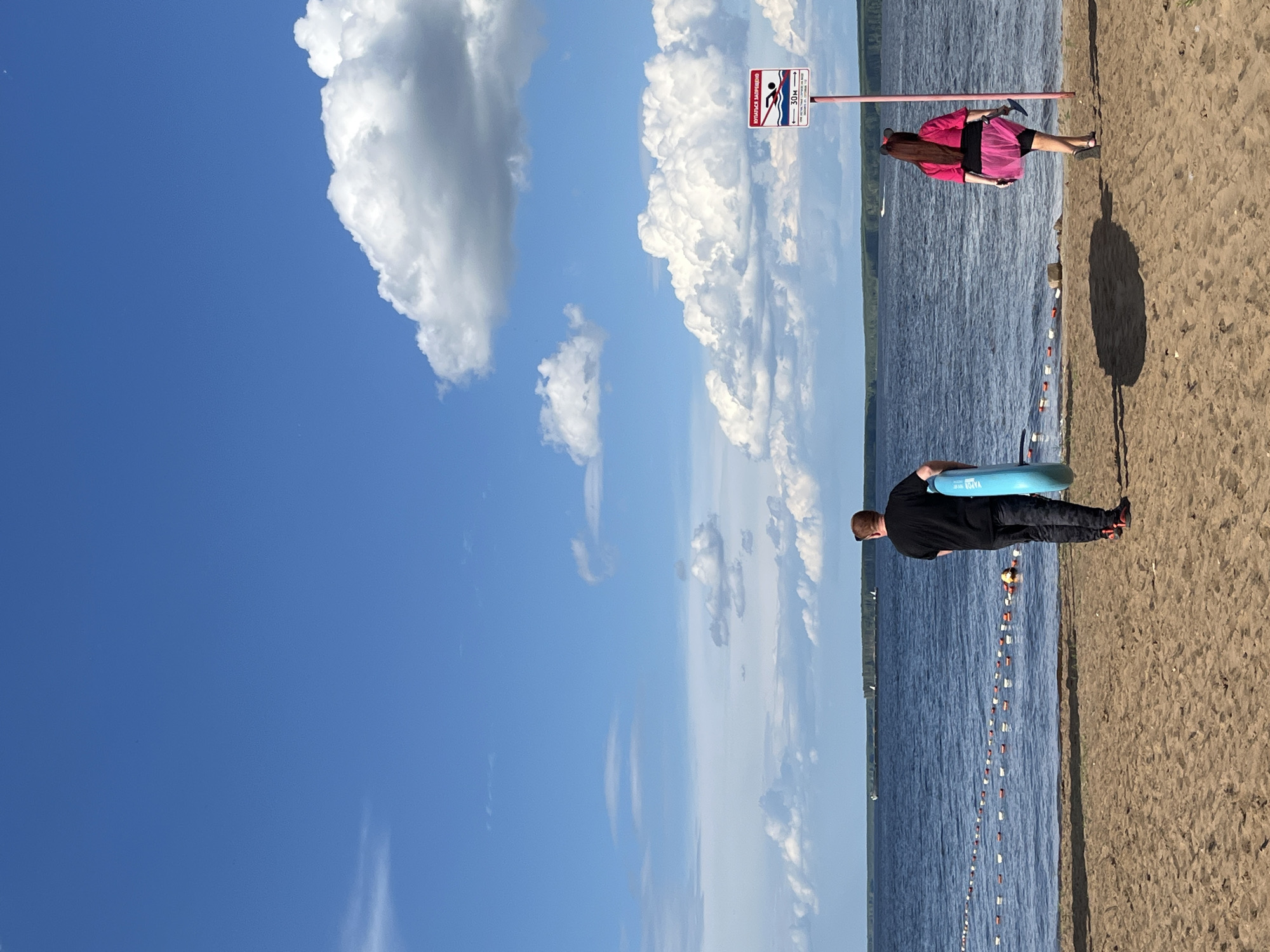 The image depicts a beach scene in the city of Chaikovsky, Russia, likely on the Kama River. The foreground is a sandy beach. In the middle ground, a man in a black t-shirt and camouflage pants walks towards the water, carrying a blue inflatable ring. To his right, a woman with dark hair styled with mouse ears, wearing a pink top and a pink skirt over black shorts, walks away from the water, holding a dark object in her right hand. A red and white sign with an image of a swimmer and text indicating swimming is prohibited is visible to the right of the woman. The background features a large body of blue water with buoys and a rope demarcating a swimming area. A tree line forms the distant horizon, and a bright blue sky with scattered white clouds fills the upper portion of the image.